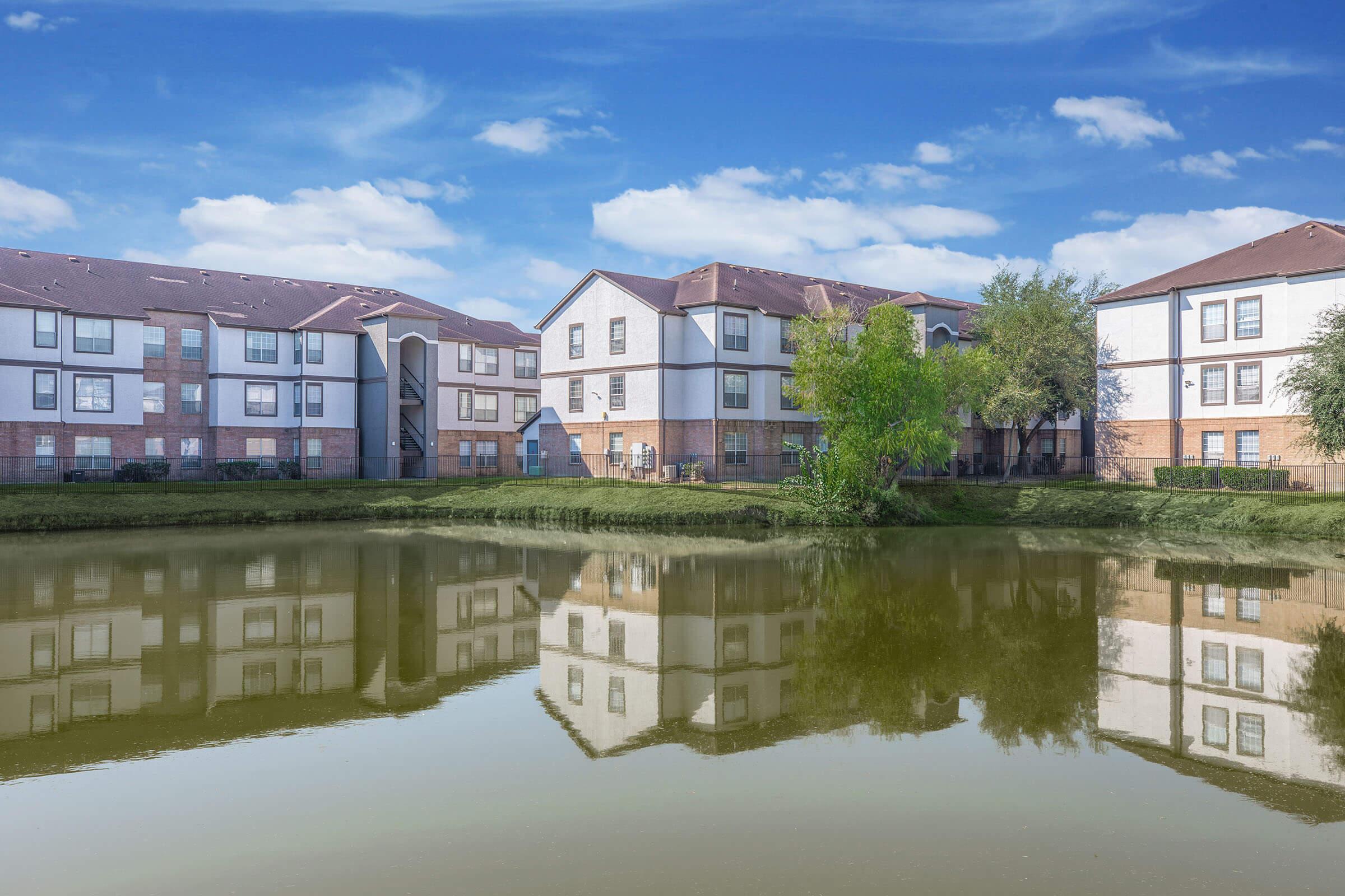 A tranquil scene featuring several multi-story apartment buildings reflected on the calm surface of a pond. The buildings are surrounded by greenery and set against a clear blue sky with scattered clouds, creating a peaceful residential atmosphere.