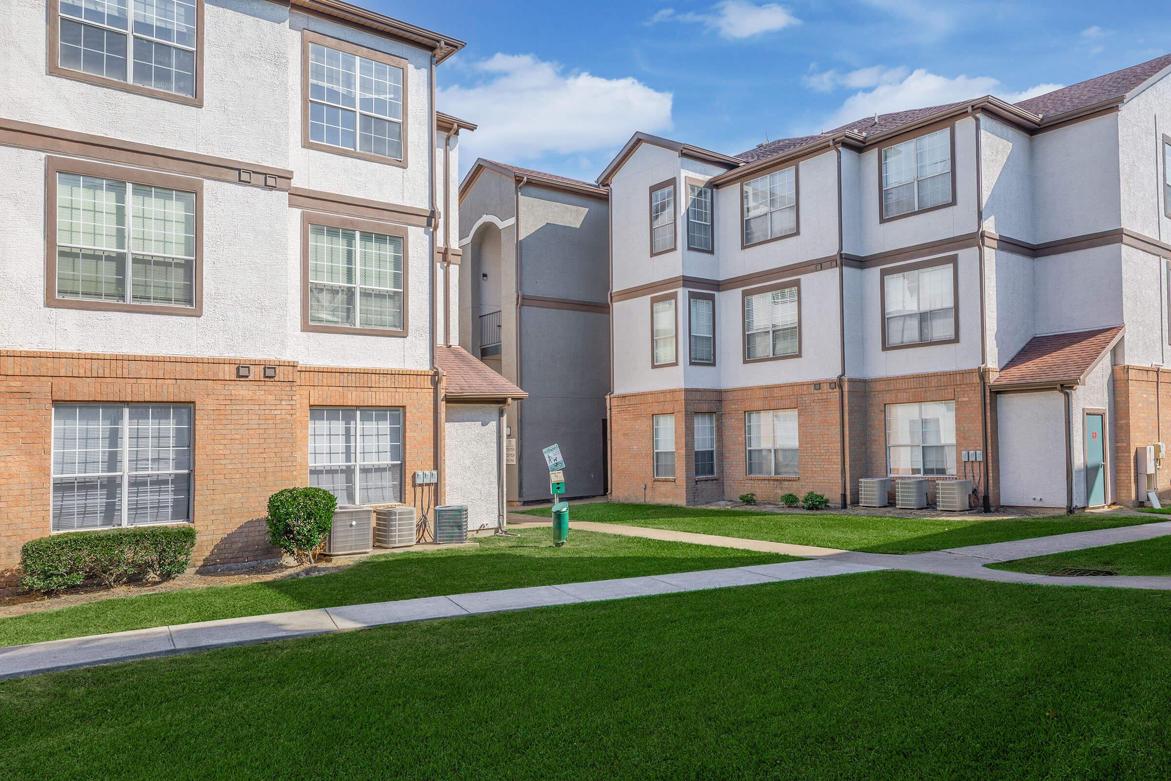 A residential apartment complex featuring multiple two-story buildings with brick and stucco exteriors. The landscape includes well-maintained grass, shrubs, and air conditioning units. Clear blue sky with scattered clouds is visible in the background.