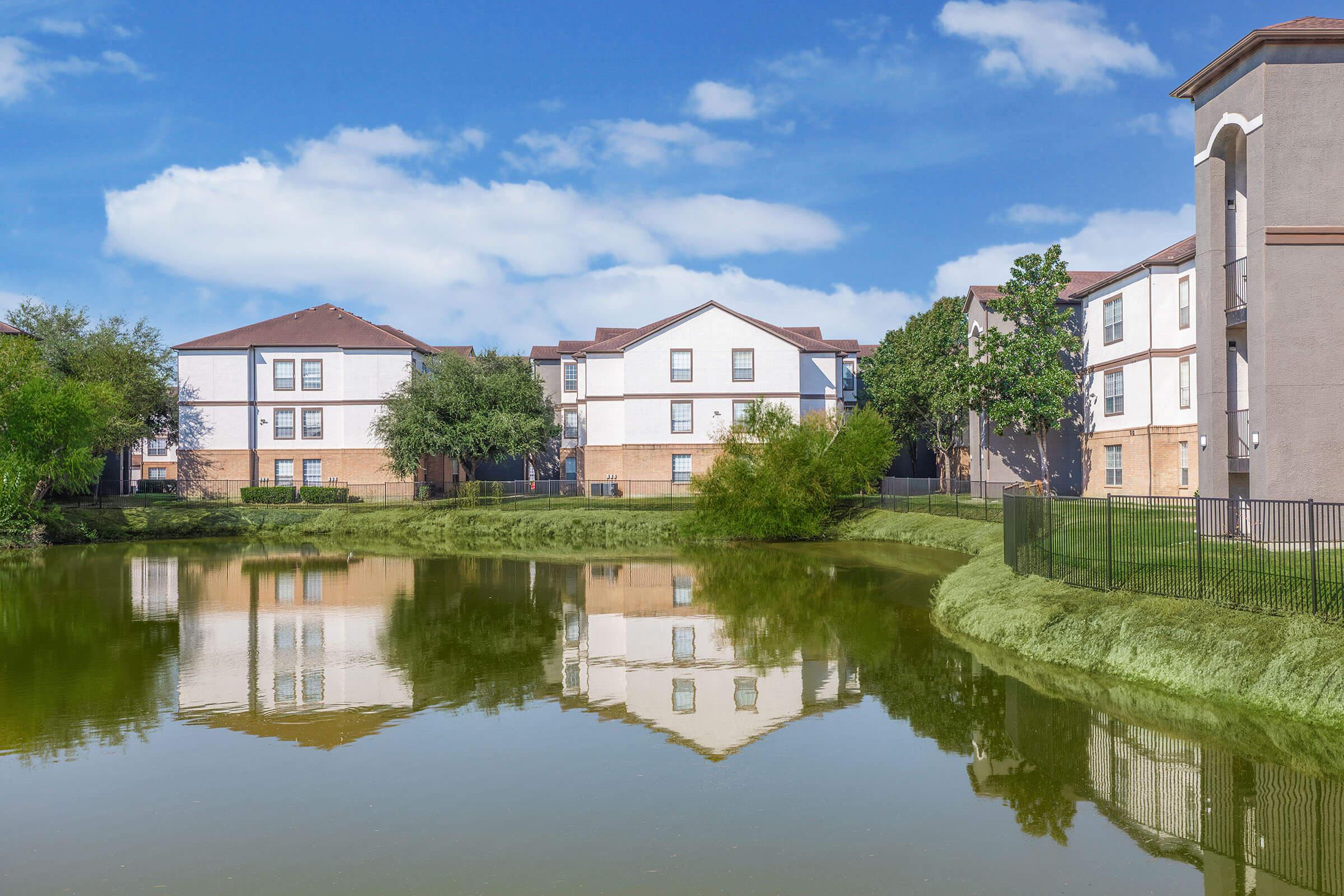 A tranquil pond reflecting white apartment buildings and lush greenery under a clear blue sky, with a few scattered clouds. The scene conveys a serene residential neighborhood environment.