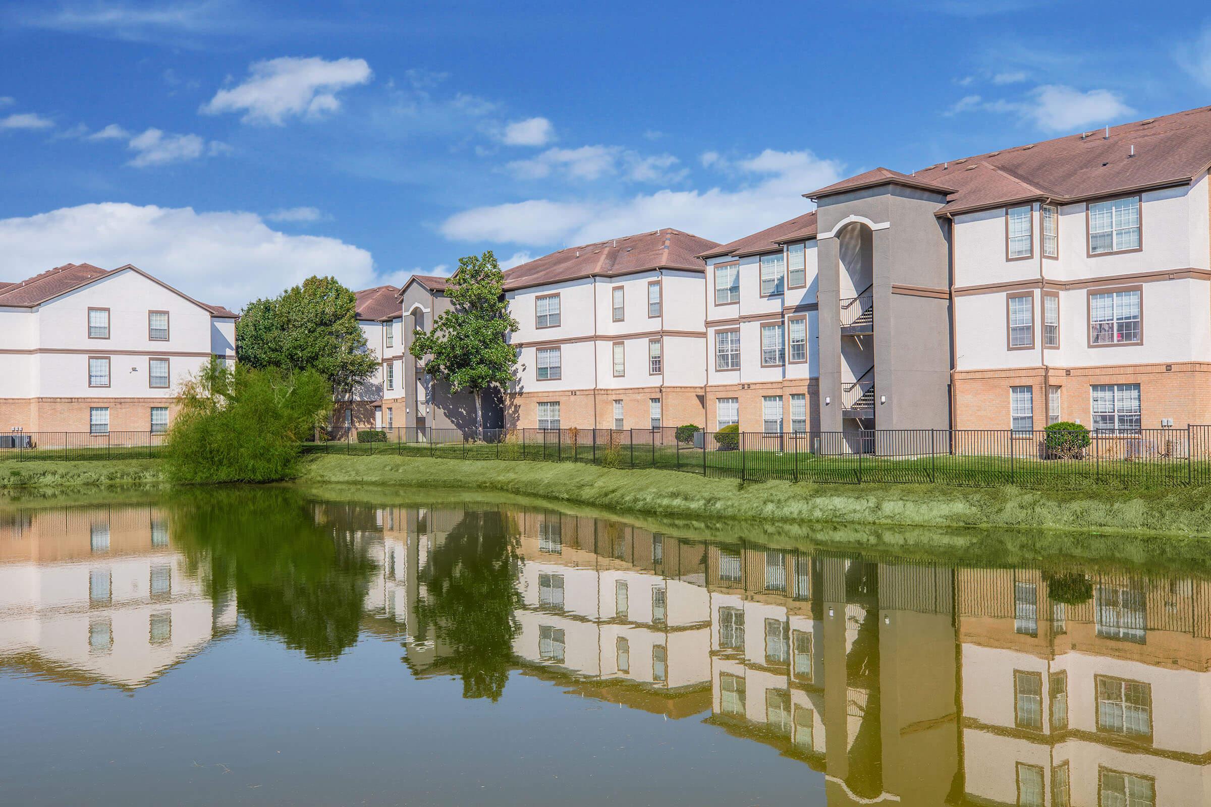A peaceful lake scene with a reflective water surface, bordered by lush green grass. On one side, several modern apartment buildings with beige and white facades are visible, under a bright blue sky with a few scattered clouds. The area is well-maintained and appears inviting.
