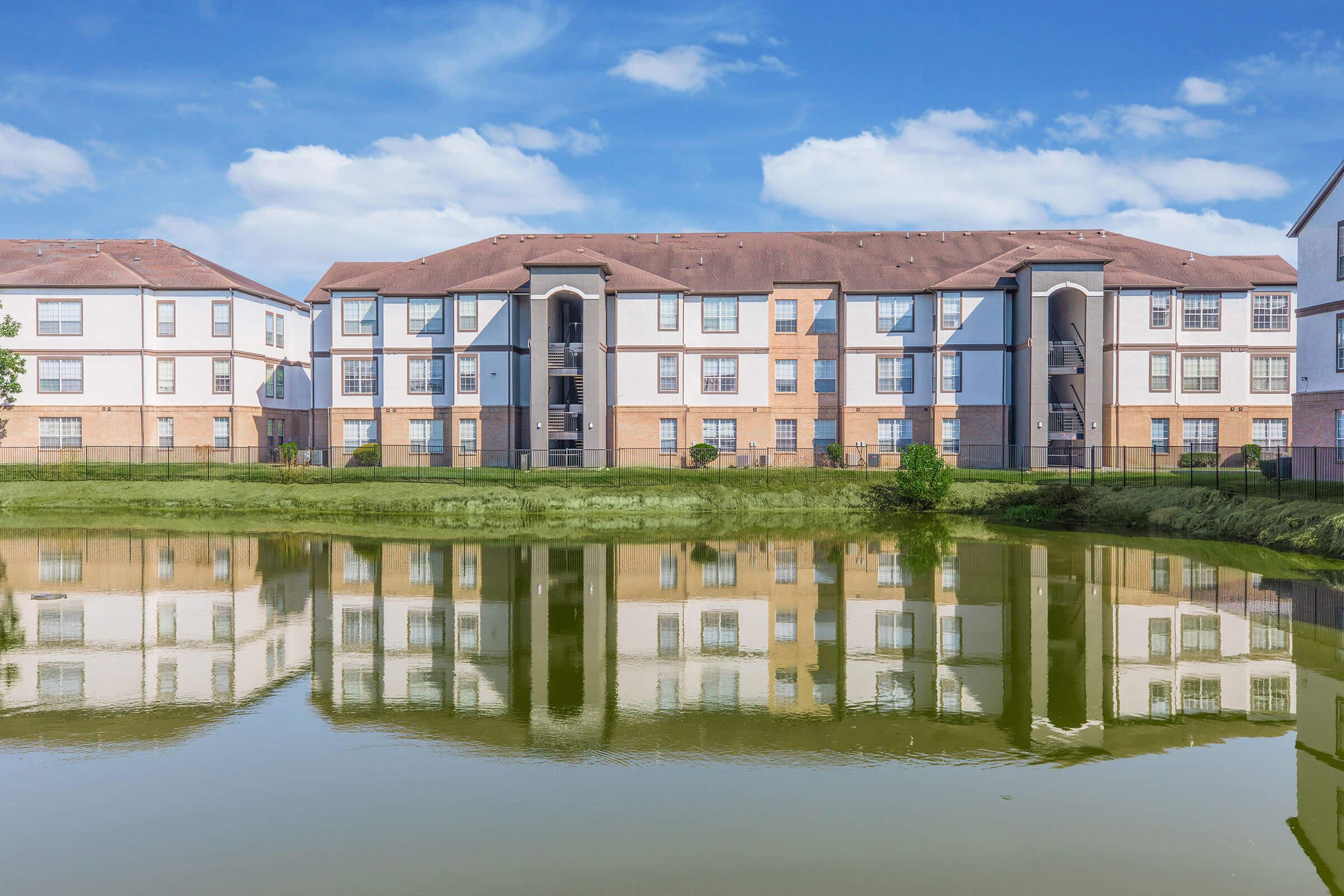 A suburban apartment complex with multiple buildings, featuring modern architectural design, is reflected in a calm pond in the foreground. The sky is blue with a few clouds, and greenery surrounds the area, creating a serene atmosphere.