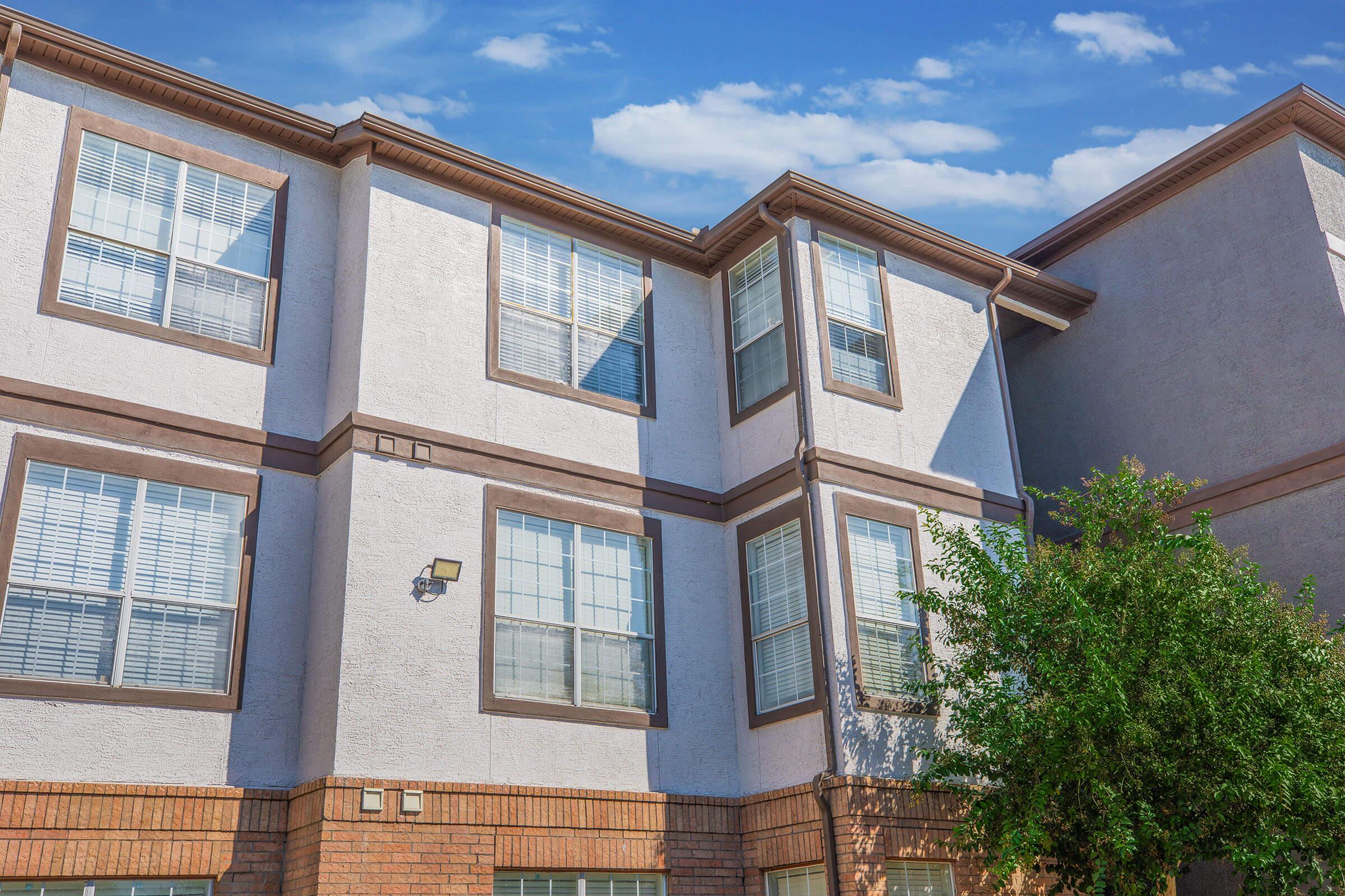 A two-story apartment building featuring a beige and brown color scheme, with multiple windows adorned with white blinds. The sky above is partly cloudy, and there is greenery at the base of the building, adding to the residential atmosphere.