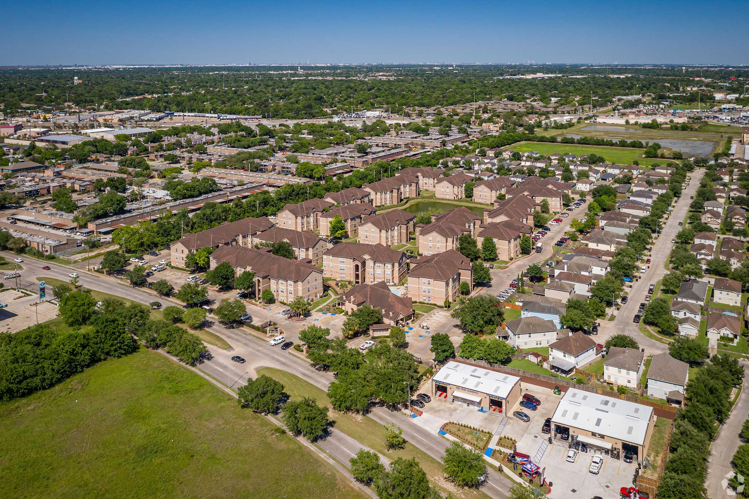 Aerial view of a residential area featuring several apartment buildings, surrounded by green lawns and trees. Nearby, there are small commercial buildings and a street lined with parked cars. In the background, cityscape and open fields are visible under a clear blue sky.