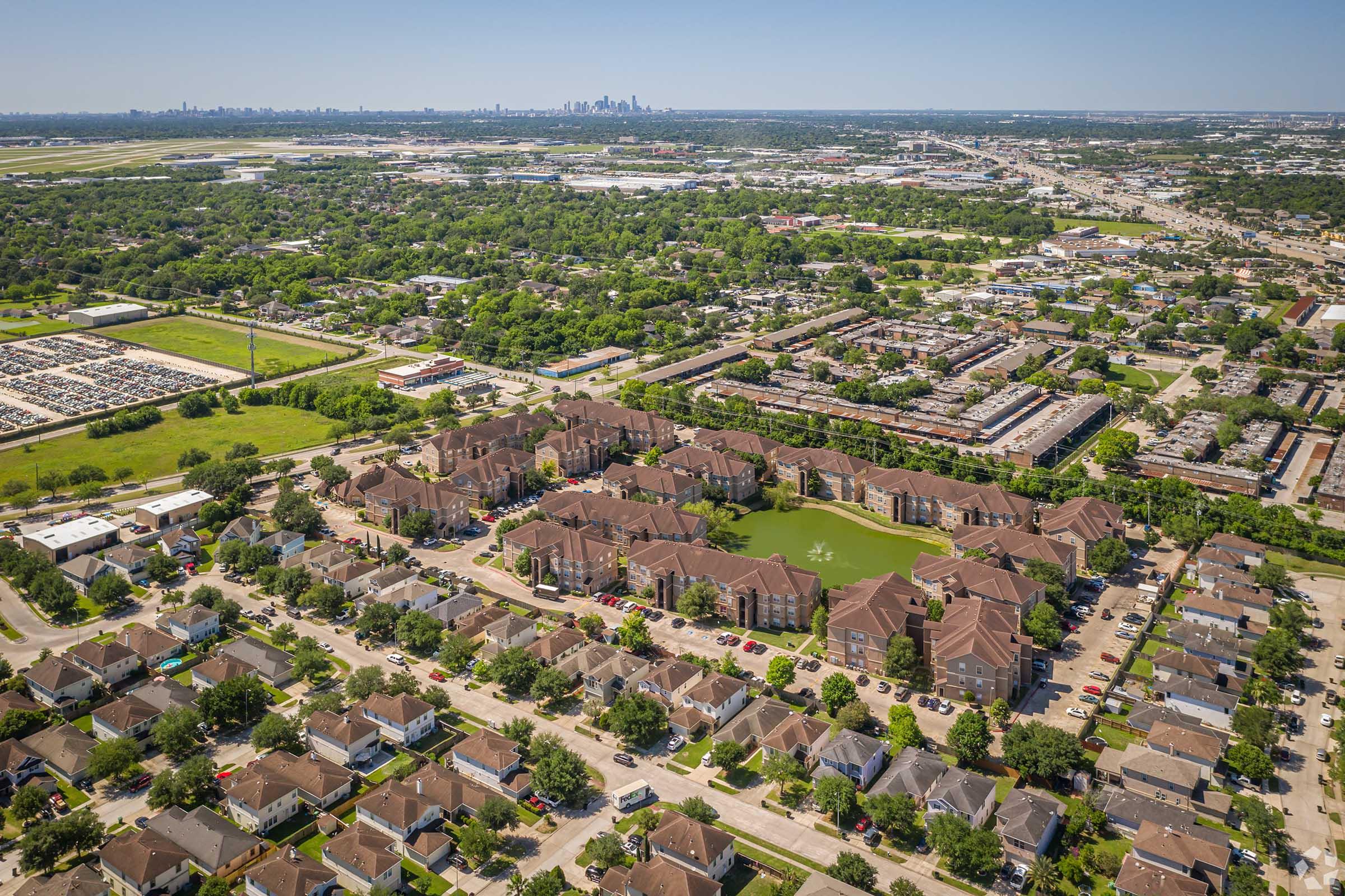 Aerial view of a residential area featuring clusters of homes and apartment buildings surrounded by greenery and a pond. In the distance, a skyline of a city can be seen along with a parking lot and open fields. The scene illustrates a mix of urban and suburban landscapes under a clear blue sky.