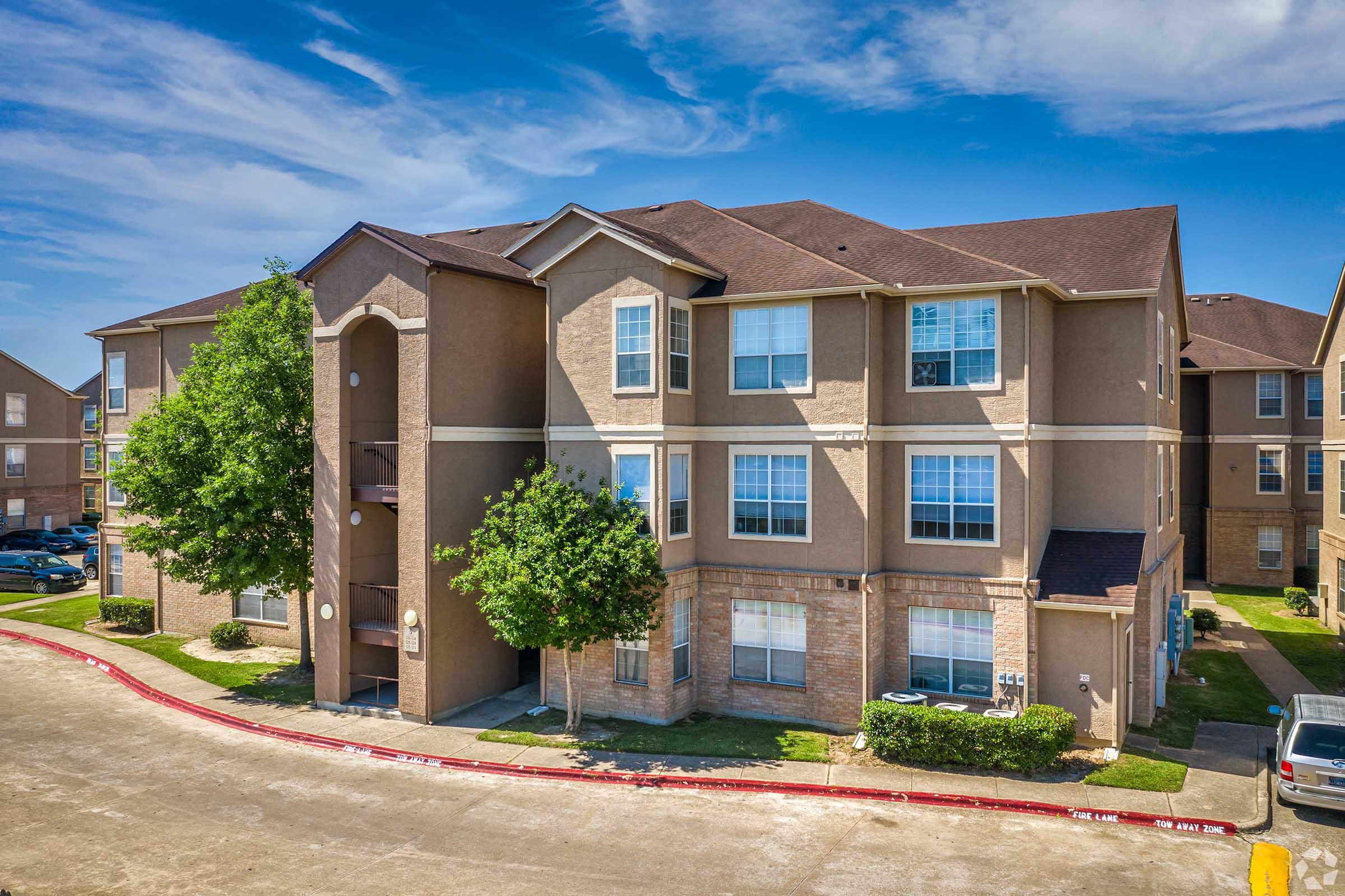 A three-story apartment building with a tan exterior and brown roofing. The building features multiple windows and small trees in front. A paved driveway runs alongside the building, and there are cars parked along the street. The sky is clear with some fluffy clouds in the background.