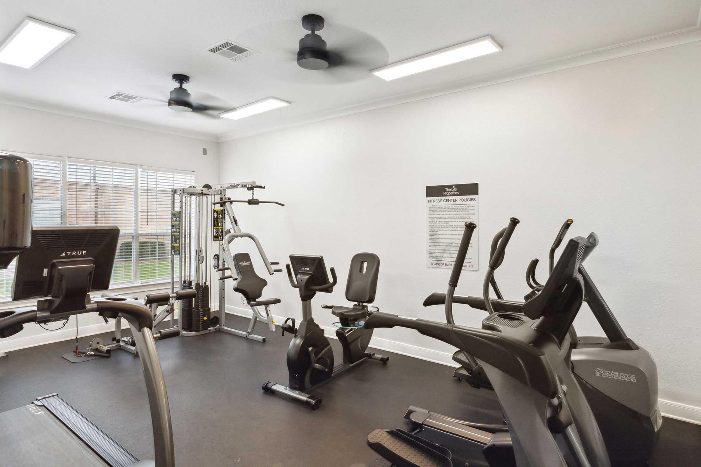 A bright and modern fitness room featuring various exercise equipment, including treadmills, stationary bikes, and a multi-station gym. Large windows allow natural light to fill the space, enhancing the inviting atmosphere. A wall board provides fitness guidelines or instructions.