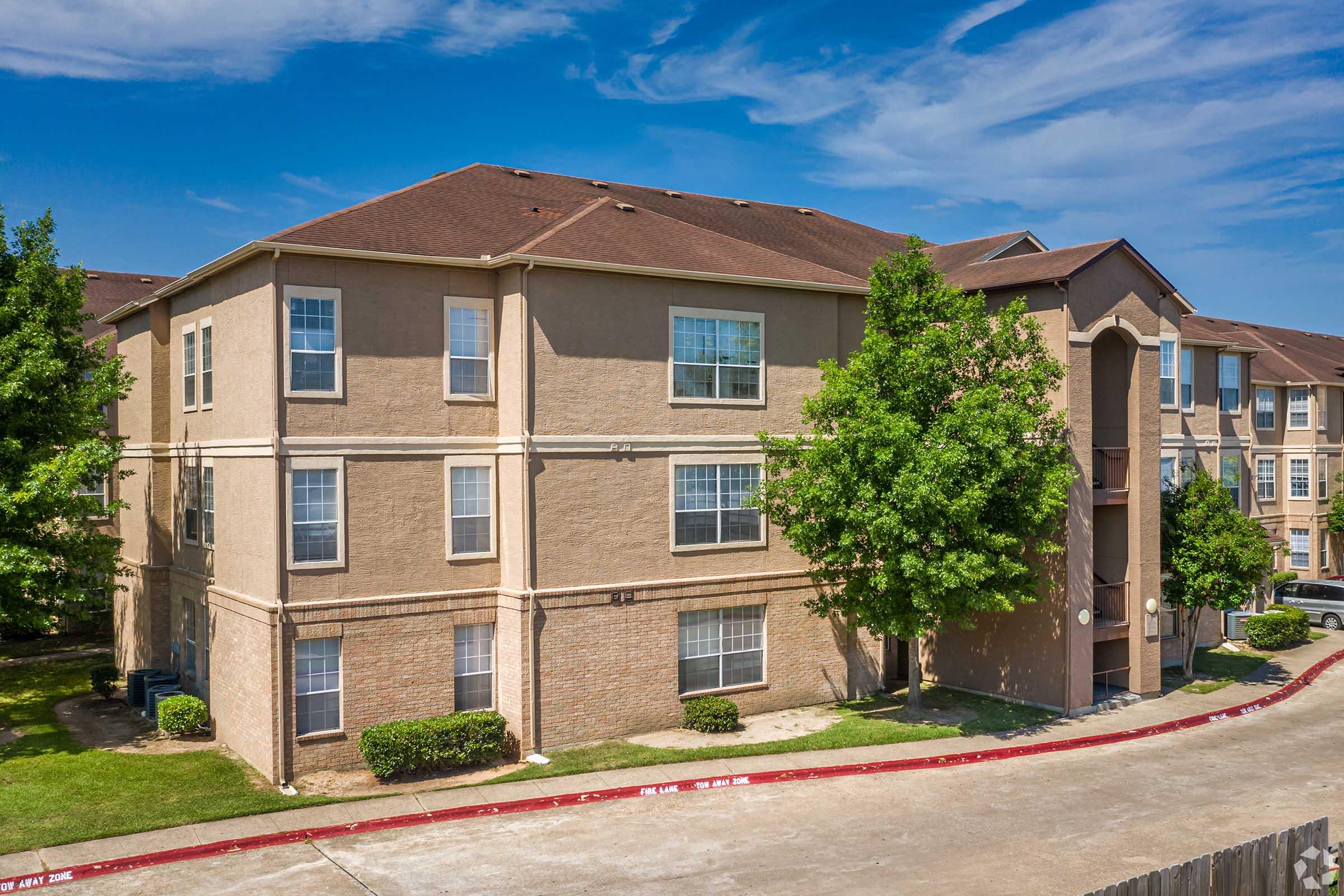 A two-story beige apartment building with multiple windows and a red curb in front. Surrounding the building are neatly trimmed shrubs and small trees. The sky is clear with some scattered clouds. The setting conveys a spacious and well-maintained residential area.