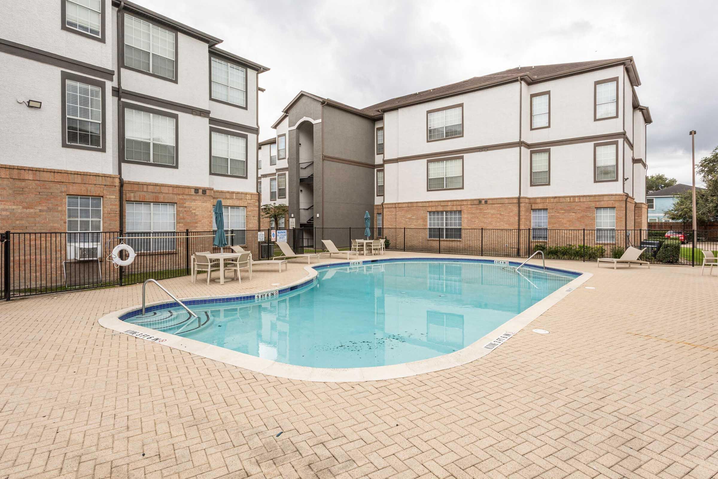 A sparkling blue swimming pool surrounded by lounge chairs and umbrellas, situated in a modern apartment complex with brick and stucco buildings. The area is enclosed with a fence, and there are trees and landscaping in the background under a cloudy sky.