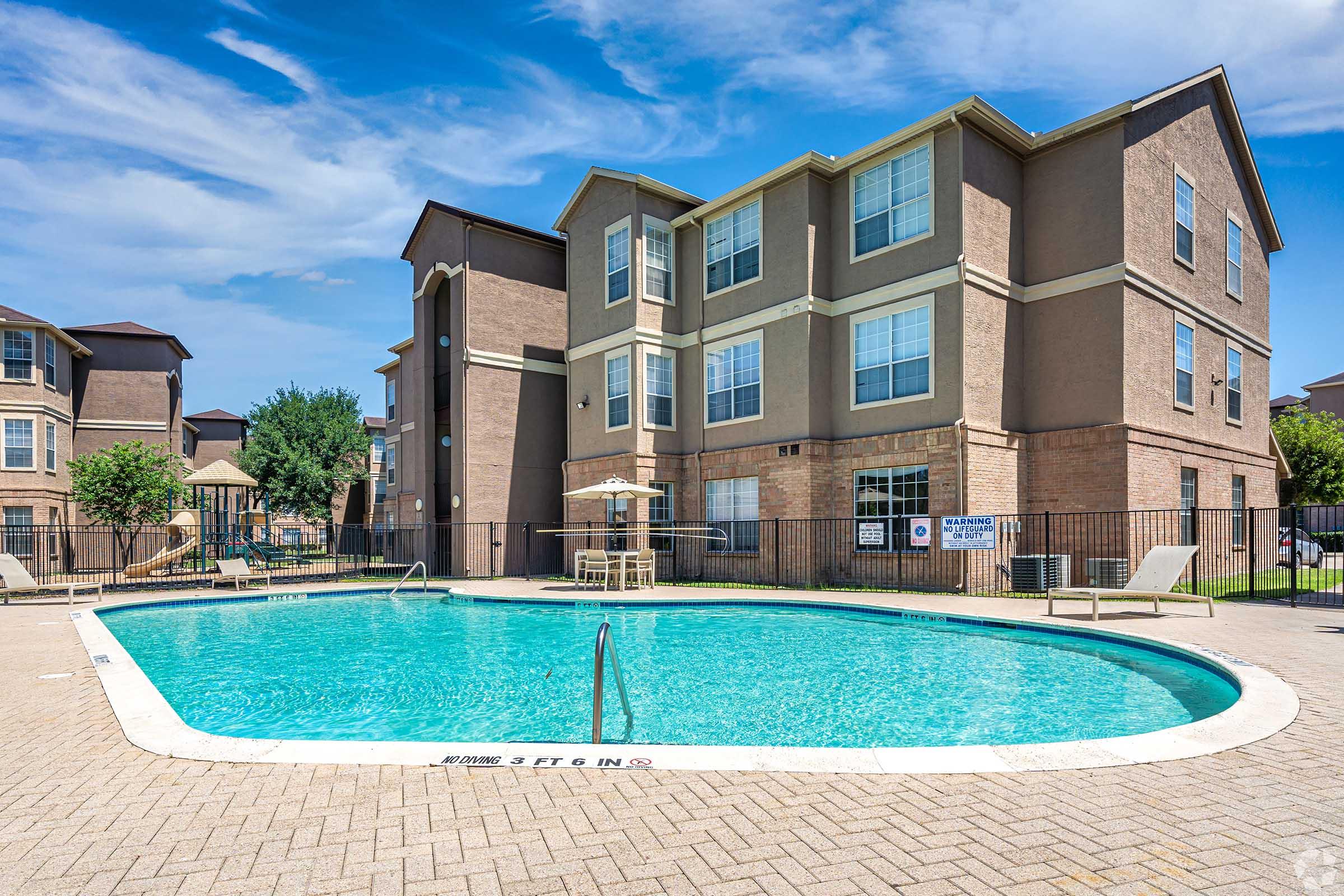 A clear blue swimming pool surrounded by a paved area and lounge chairs, located at an apartment complex. The two-story brick building in the background features large windows and a well-maintained exterior. The sky is partly cloudy, adding to the inviting atmosphere of the pool area.