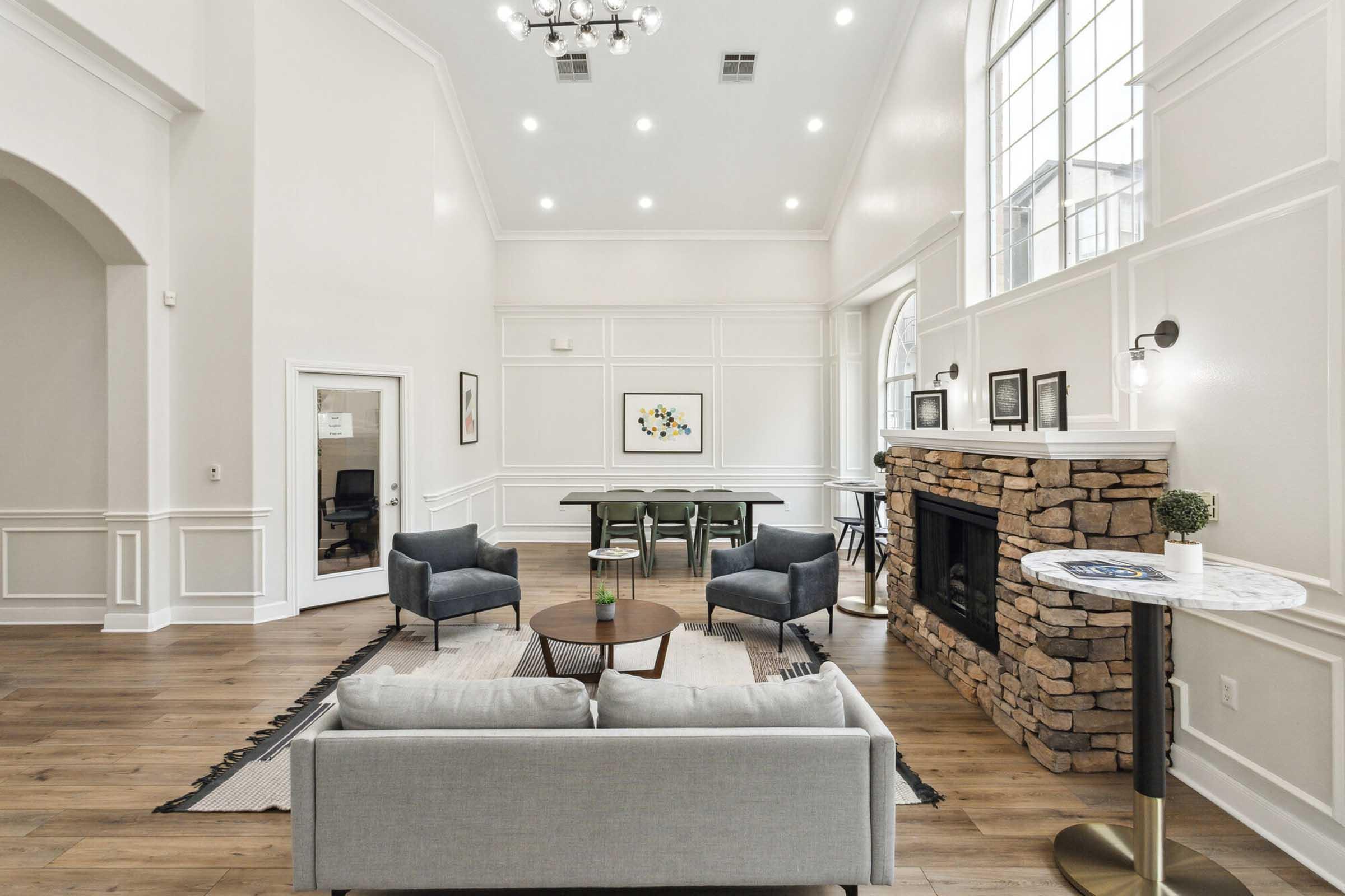A spacious, modern living area featuring a stone fireplace, comfortable gray sofas, and two armchairs. Large windows let in natural light, complemented by stylish lighting fixtures. A dining table with green chairs is visible in the background, enhancing the inviting atmosphere.