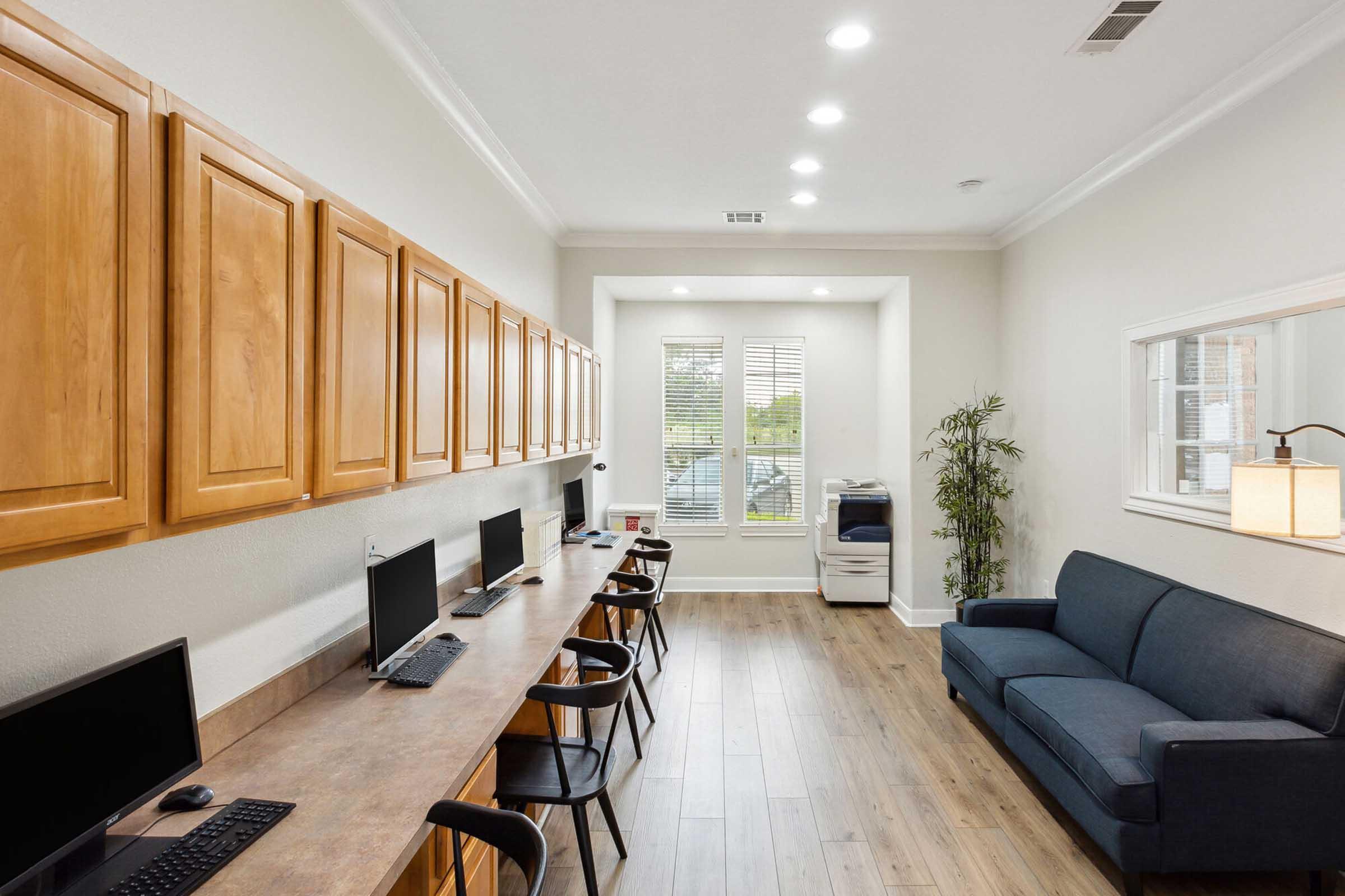 A modern office space featuring light-colored walls and wooden cabinetry. Several computers are set up on a long wooden desk, with black chairs. A blue sofa is positioned against the wall, and a potted plant is nearby. Natural light comes in through a window, creating a bright and inviting atmosphere.
