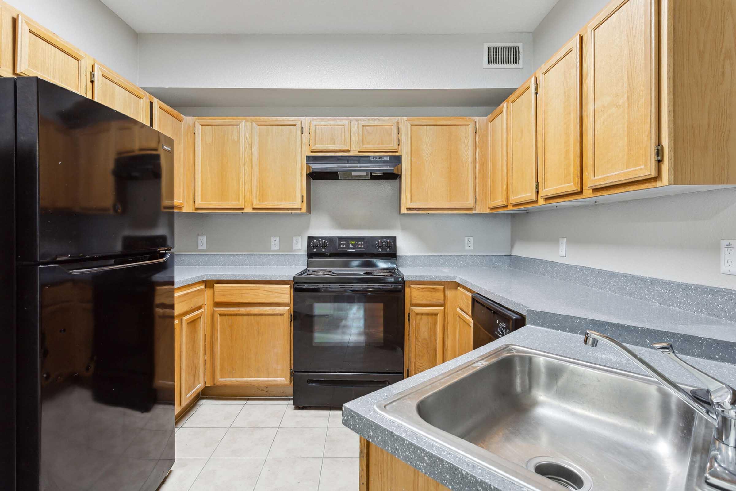 Kitchen featuring wooden cabinets, a black refrigerator, and a black stove with a vent above. The countertops are gray, and a stainless steel sink is visible in the foreground. The walls are painted a light color, providing a simple and clean aesthetic.