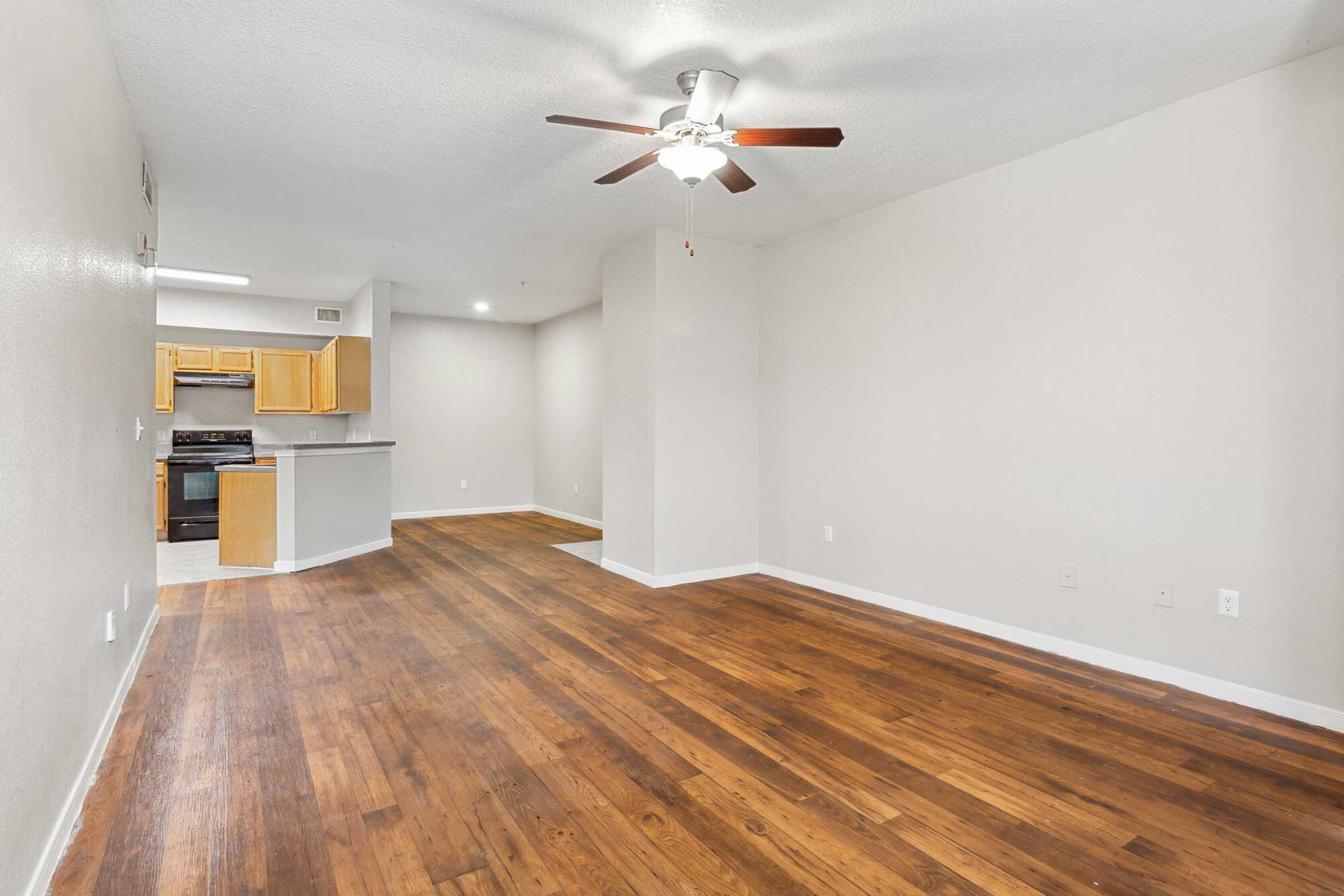 A spacious, empty living room with hardwood floors and a ceiling fan. To the left, there is an open kitchen featuring wooden cabinets and modern appliances. The walls are painted a light color, contributing to a bright and airy atmosphere. Natural light enters the space, enhancing the inviting ambiance.