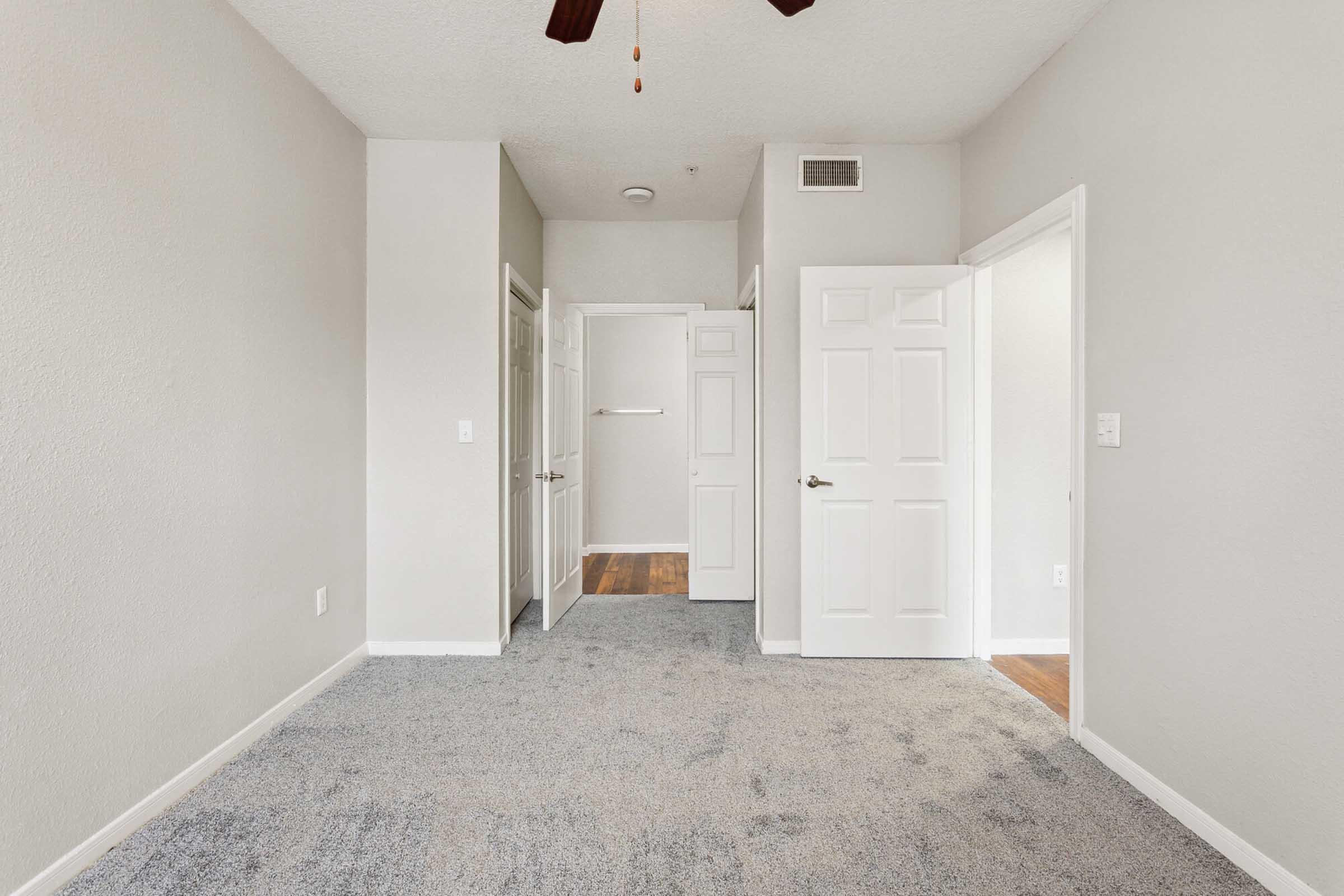 A view of a light-colored room with carpeted flooring. There are two closed doors on the left leading to small closets and a third open door on the right leading to another room. A ceiling fan is visible above, and the walls are painted in a neutral tone. The overall appearance is clean and well-lit.