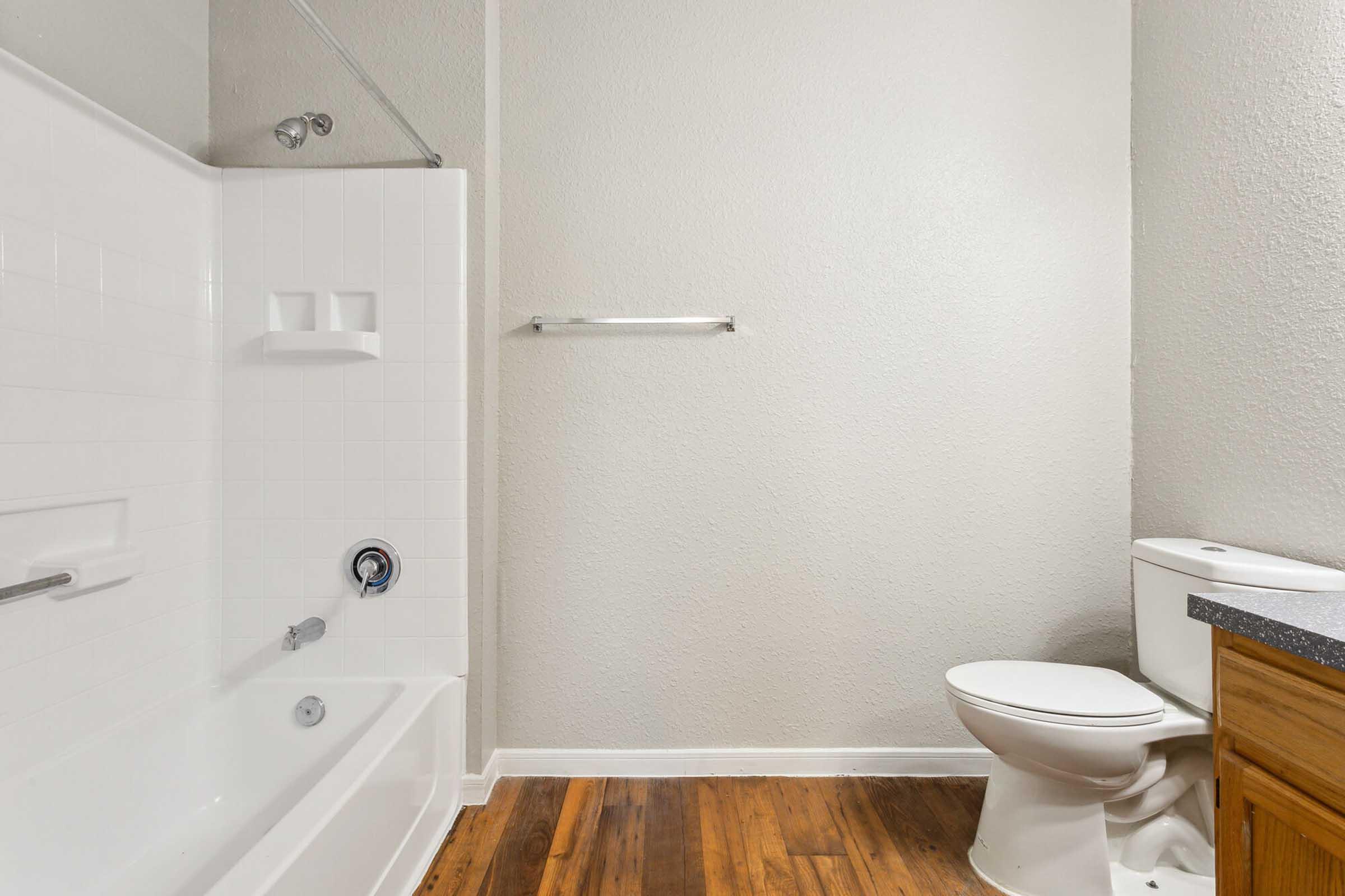 A clean and modern bathroom featuring a white bathtub with a showerhead, a toilet, and a wooden vanity. The walls are painted in a light color, and the floor is made of hardwood. A towel bar is mounted on the wall next to the bathtub.