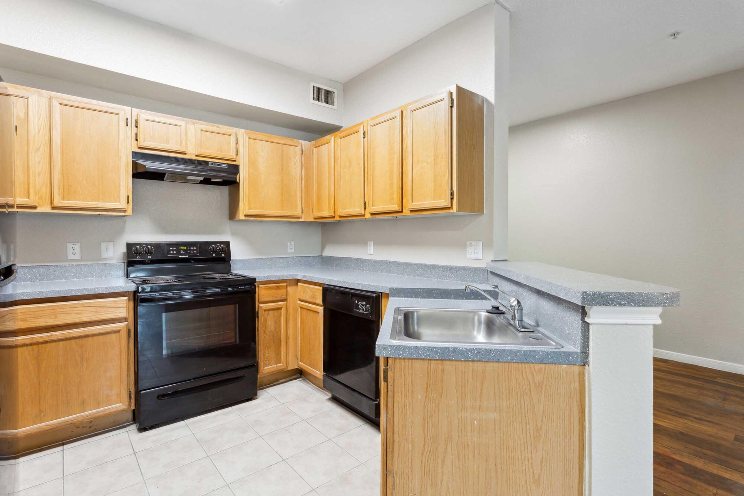 A modern kitchen featuring light wood cabinetry, a black stove and dishwasher, and gray countertops. The layout includes a sink integrated into the counter with a tiled floor. The walls are painted in a neutral shade, providing a bright and spacious feel.