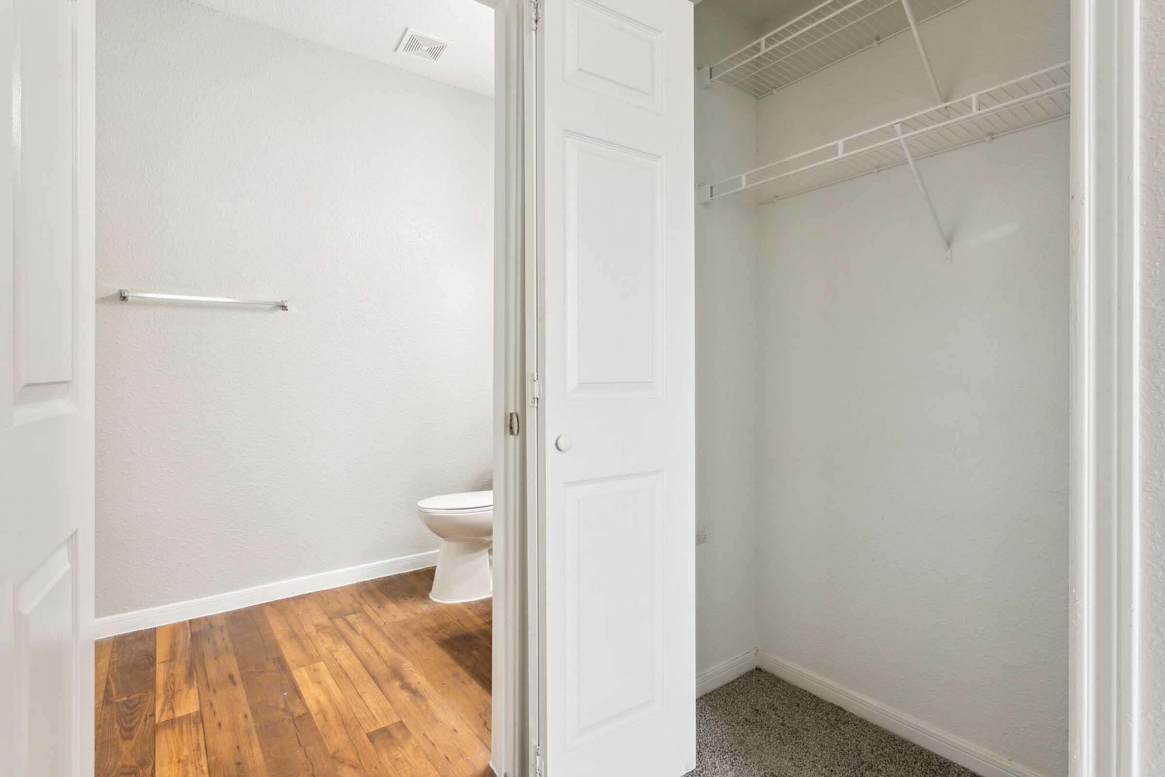 A small bathroom with light gray walls, a toilet visible in the corner, and wooden flooring that transitions to carpet at the doorway. There is a closet with an open door featuring a wire shelf. A towel rack is mounted on the wall opposite the toilet. Natural light brightens the space.