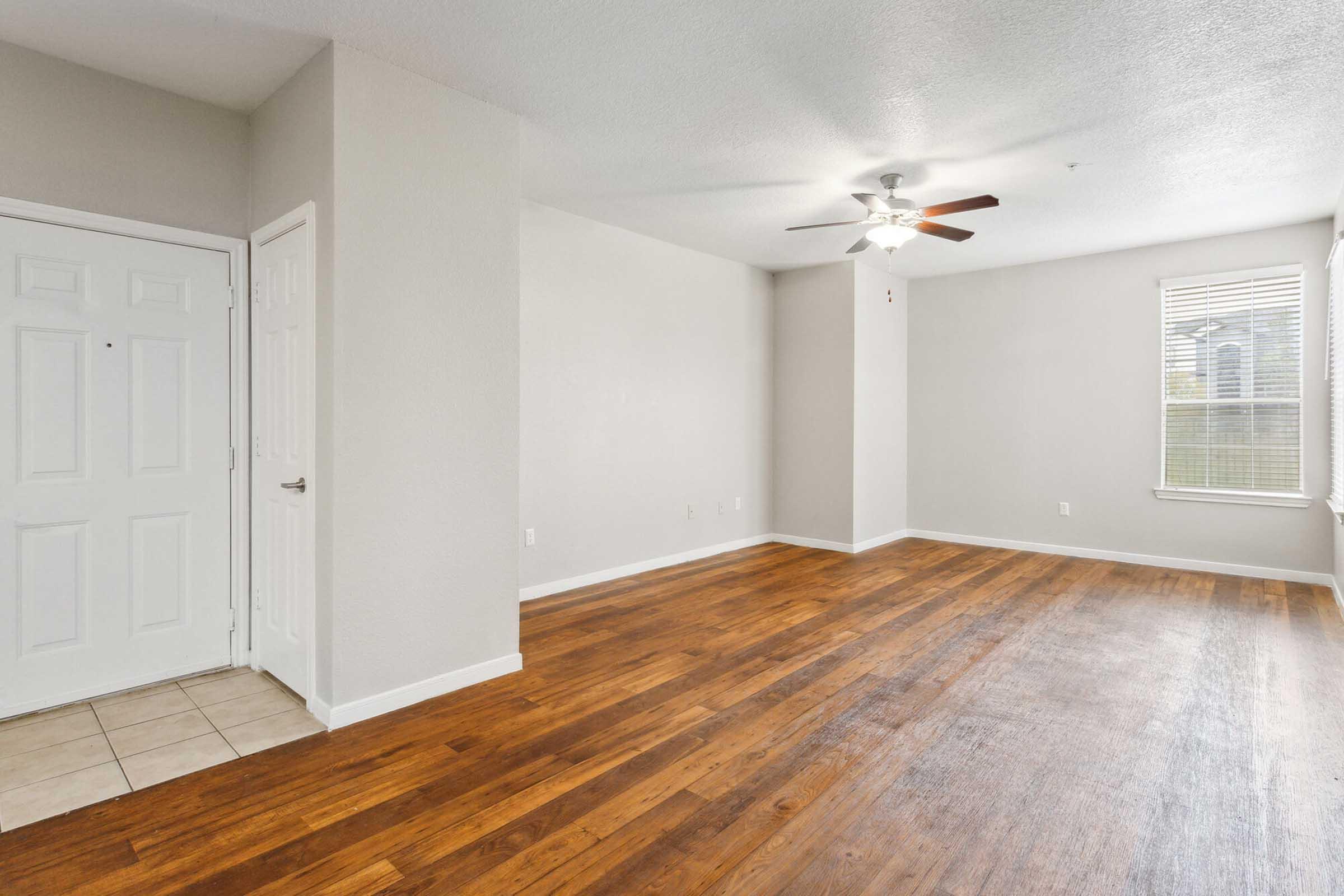 A spacious living area with hardwood floors and a ceiling fan. The walls are painted light gray, and there is a window allowing natural light. To the left, there is a white door leading to another room, and tiles are visible near the entrance.
