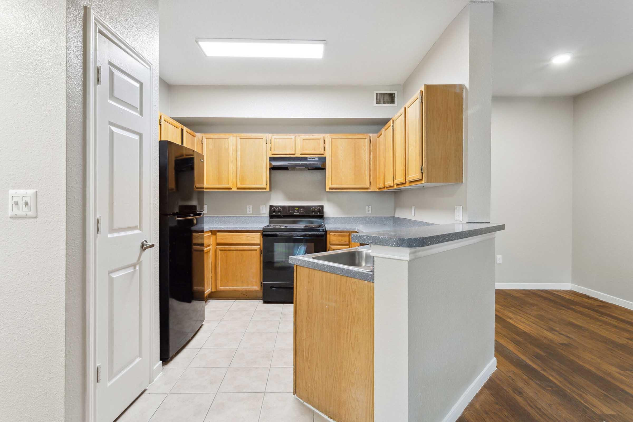 A modern kitchen featuring light wood cabinetry, black appliances, and a gray countertop. The space has beige walls and tile flooring, with an open layout leading to a living area. A white door is visible on the left side, enhancing the room's natural light from overhead lighting.