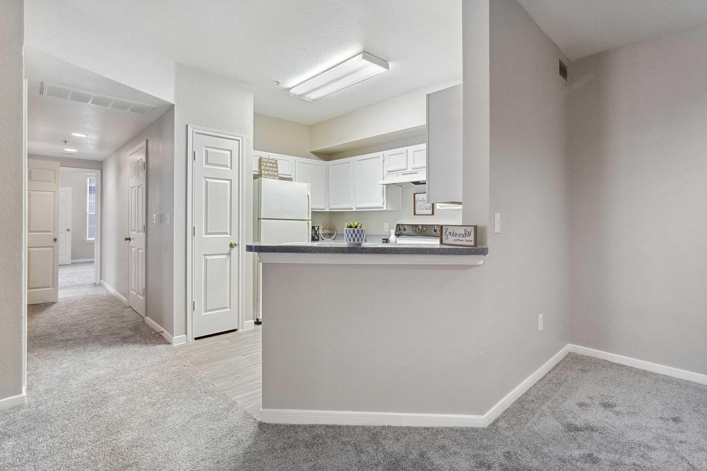 Spacious and bright interior of a modern apartment featuring a kitchen area with white cabinets, a counter bar, and a hallway with light-colored walls and plush carpeting. A refrigerator is visible in the background, along with doors leading to other rooms.