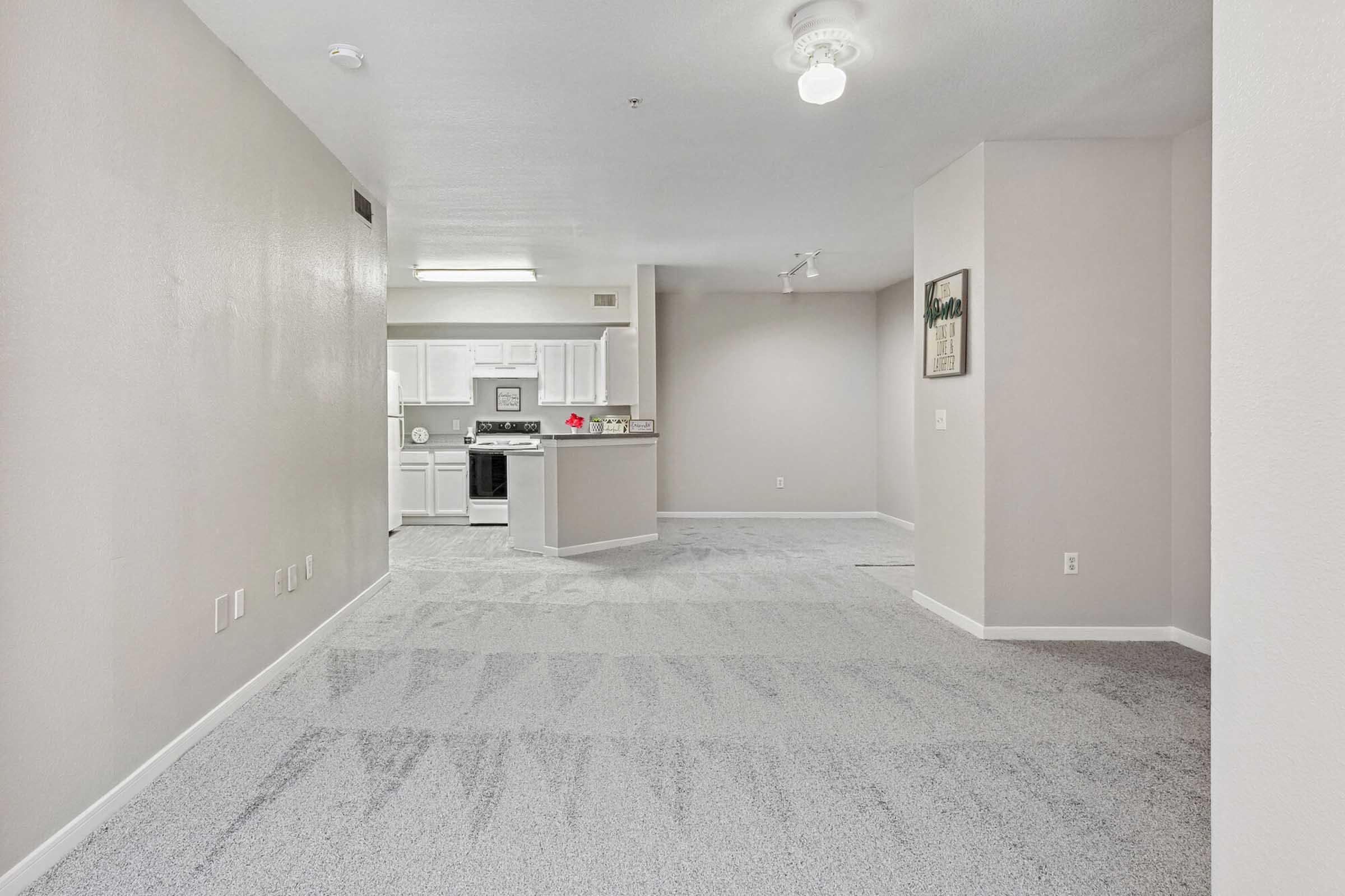 Spacious interior view of a light-colored living area with a carpeted floor. In the background, a kitchen area is visible, featuring white cabinets and appliances. The walls are painted in neutral tones, and there is a ceiling light fixture. No furniture is present, giving a sense of openness.