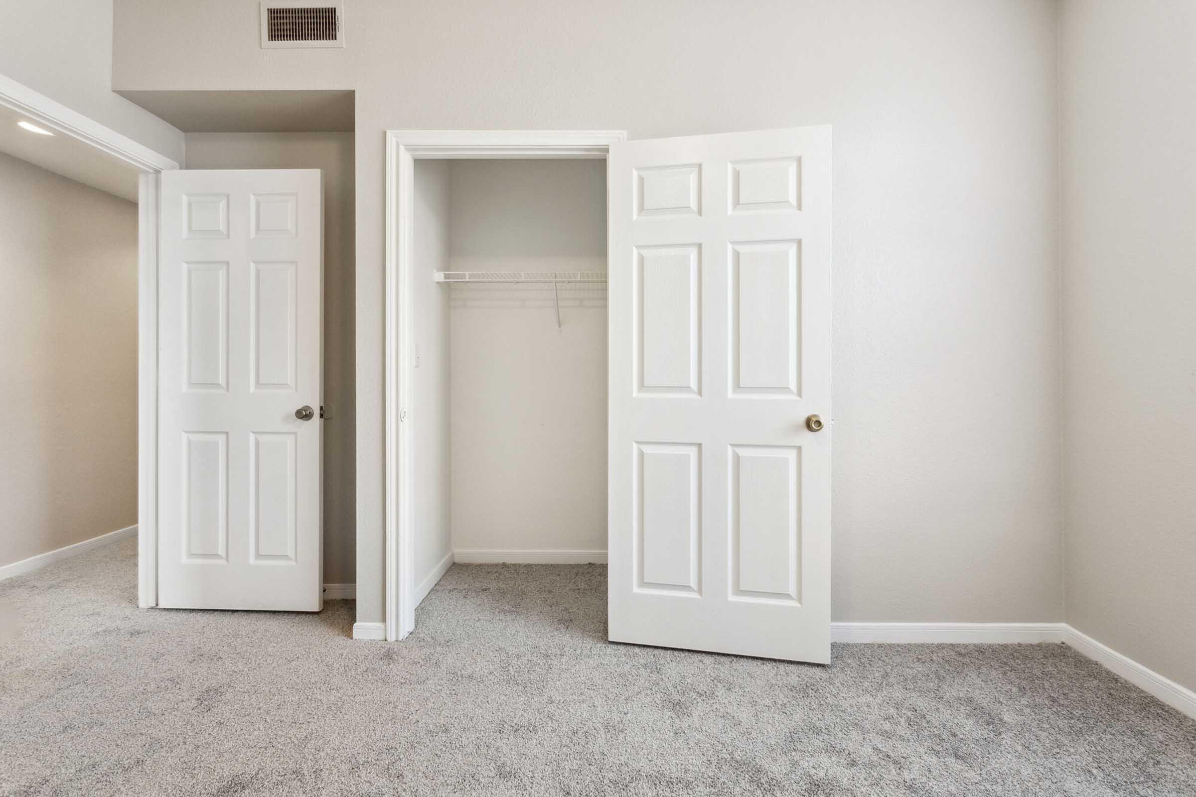 A clean, neutral-toned room featuring two open white doors leading to a closet with a hanging shelf. The floor is covered in light gray carpet, and the walls are painted in a soft shade, contributing to a spacious and inviting atmosphere.