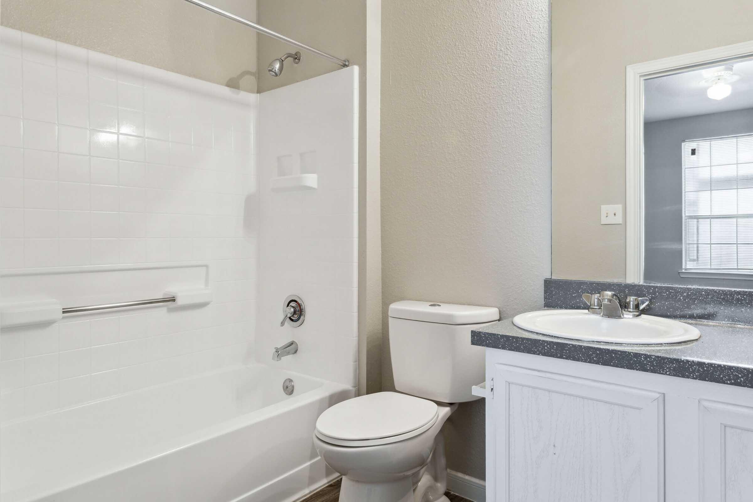 A clean and modern bathroom featuring a white bathtub with a shower, a toilet, and a sink with a gray countertop. The walls are a light beige color, and there is a mirror above the sink. Natural light enters through a window with a blind, creating a bright atmosphere.