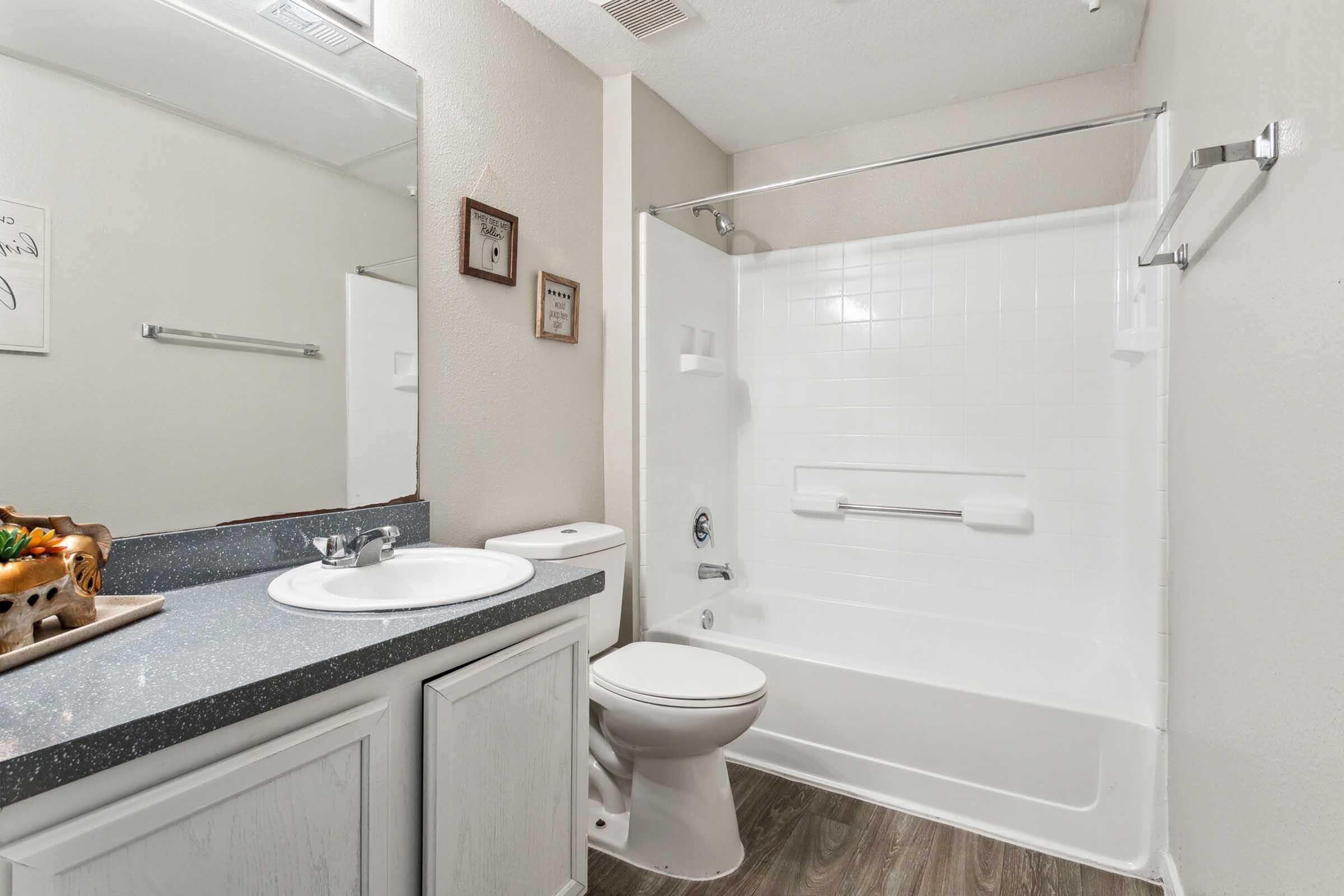 A clean and modern bathroom featuring a white bathtub with a shower, a sink with a countertop, and a toilet. The walls are painted in a light color, and there are decorative frames on the wall. The floor has a wood-like finish, adding a warm touch to the space.