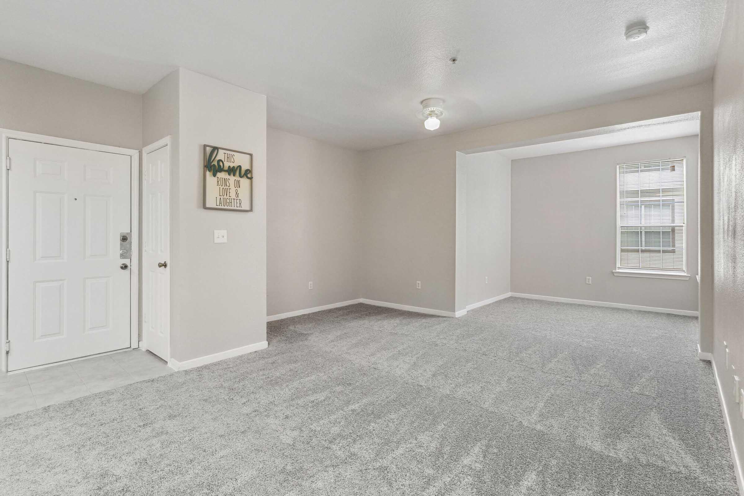Spacious interior of a residential living area featuring light gray carpet, neutral walls, a front door with a small window, and a decorative wall sign. The room is well-lit with natural light coming through a window in the background, creating a welcoming atmosphere.