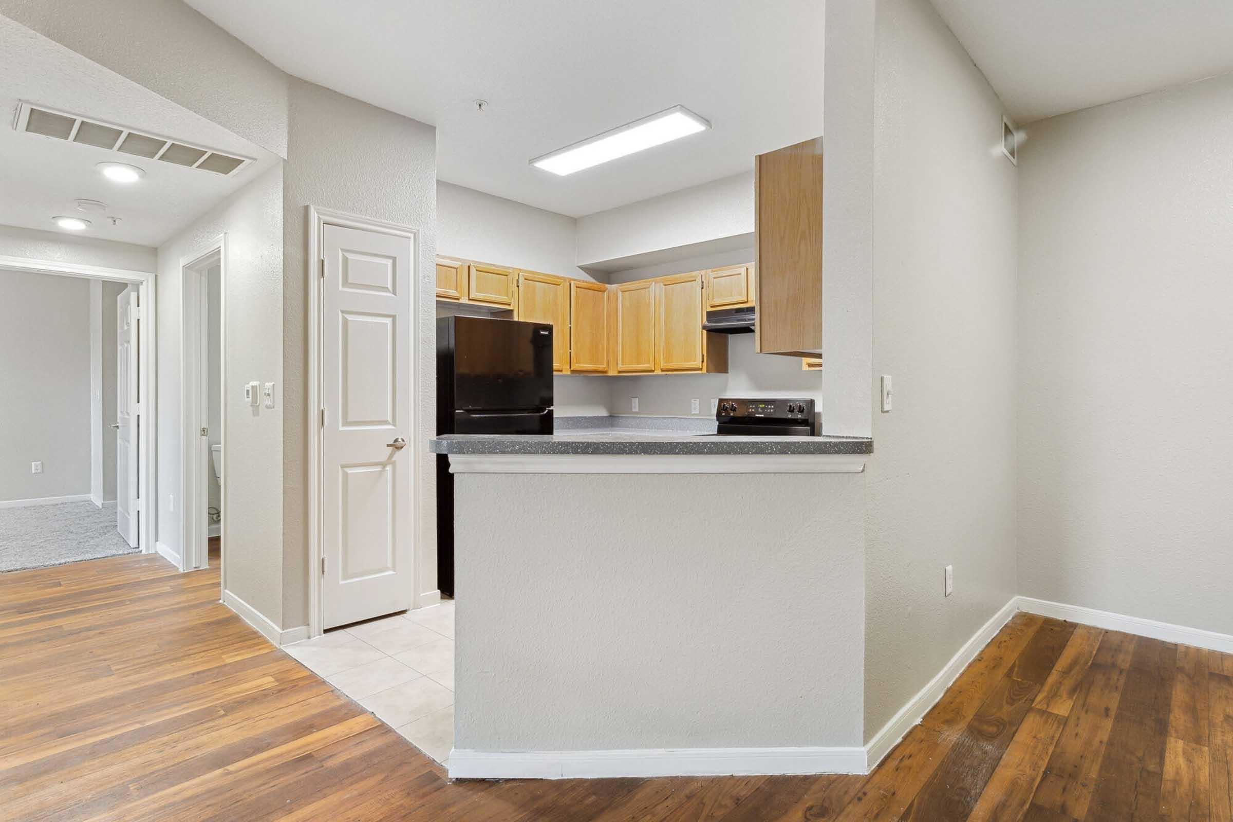 View of a modern kitchen area with wooden cabinets, black appliances, and a countertop. The open layout leads to a hallway and a carpeted area, featuring light-colored walls and a blend of tile and wooden flooring. Natural light illuminates the space adequately.