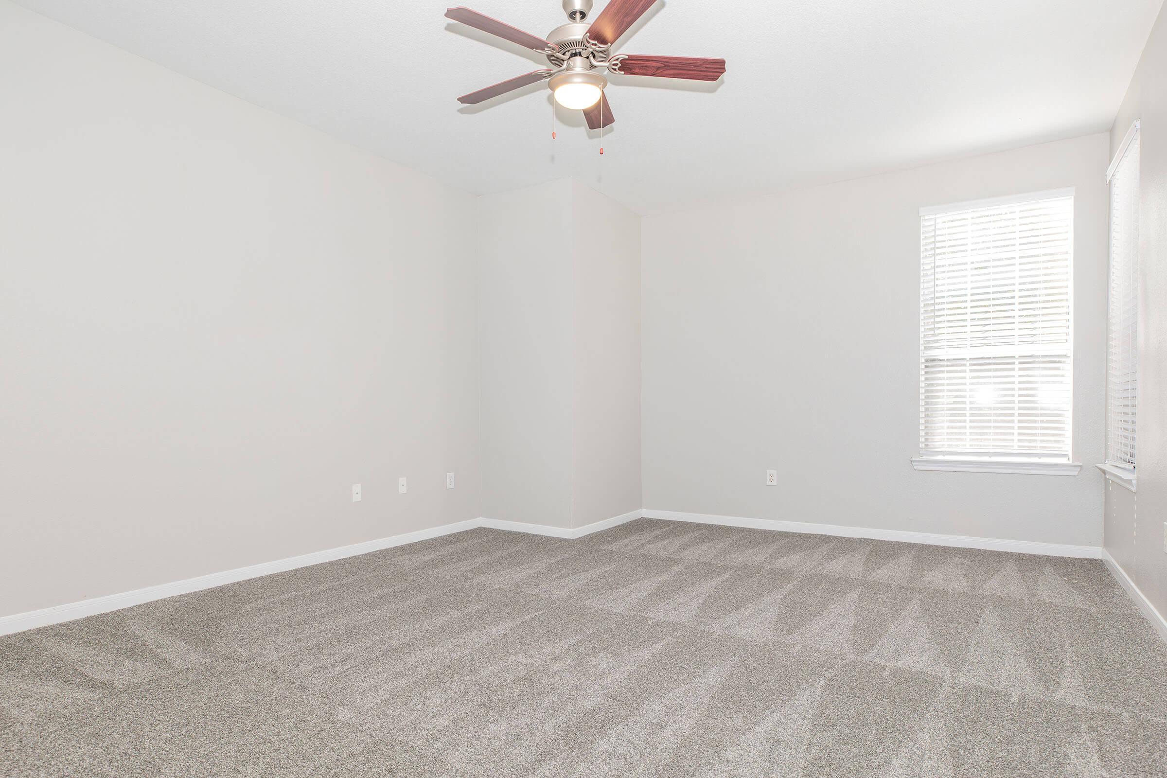A spacious, empty room featuring light gray walls and carpet. It is illuminated by natural light coming through a window with white blinds. A ceiling fan with wooden blades is centered in the room. The overall atmosphere is bright and airy, suitable for various purposes.