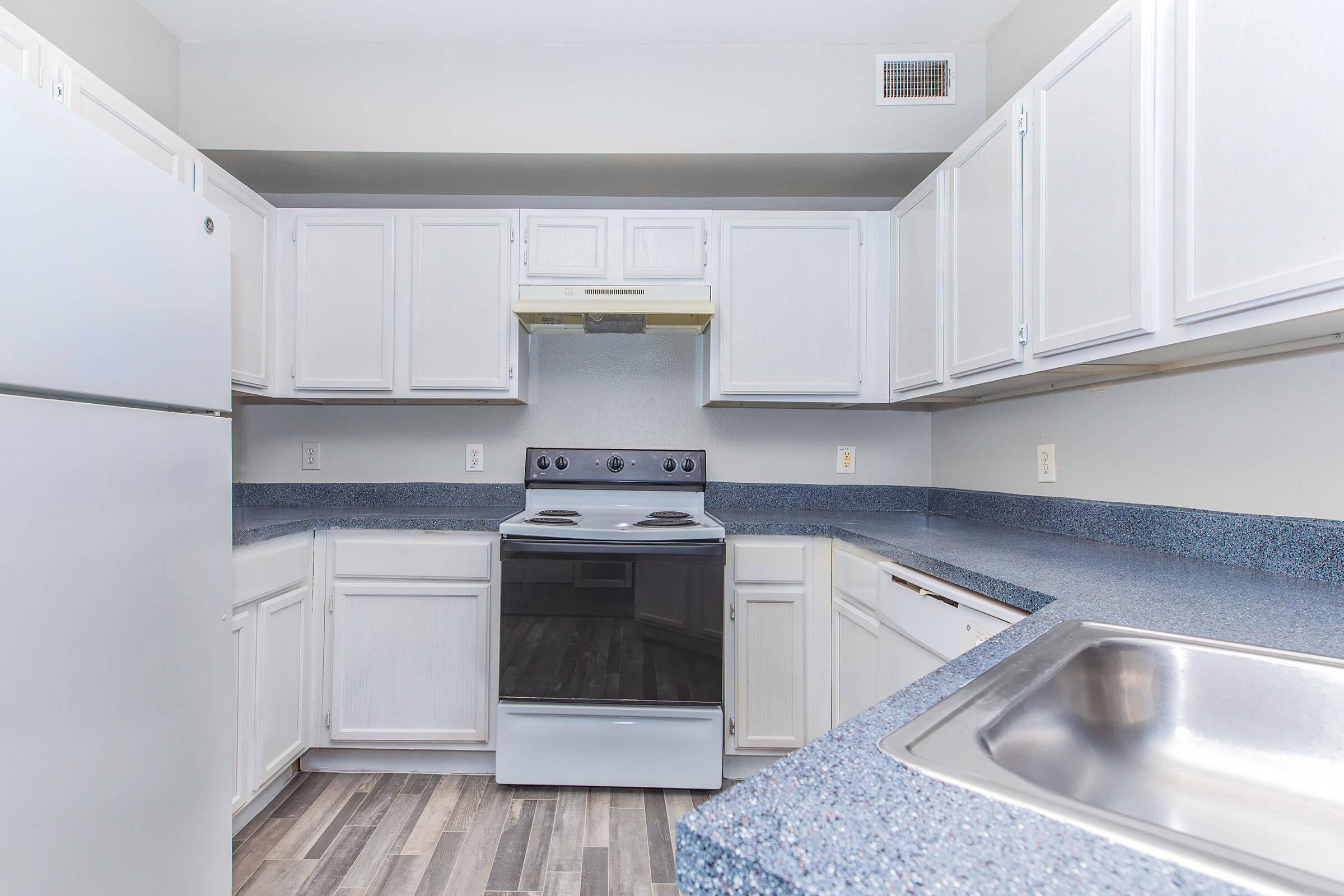 Modern kitchen featuring white cabinets, a black stove and oven, stainless steel sink, and blue countertops. The walls are painted in a light color, and the flooring is a wood-like laminate. The kitchen is well-lit with ample cabinet storage and a clean, minimalistic design.