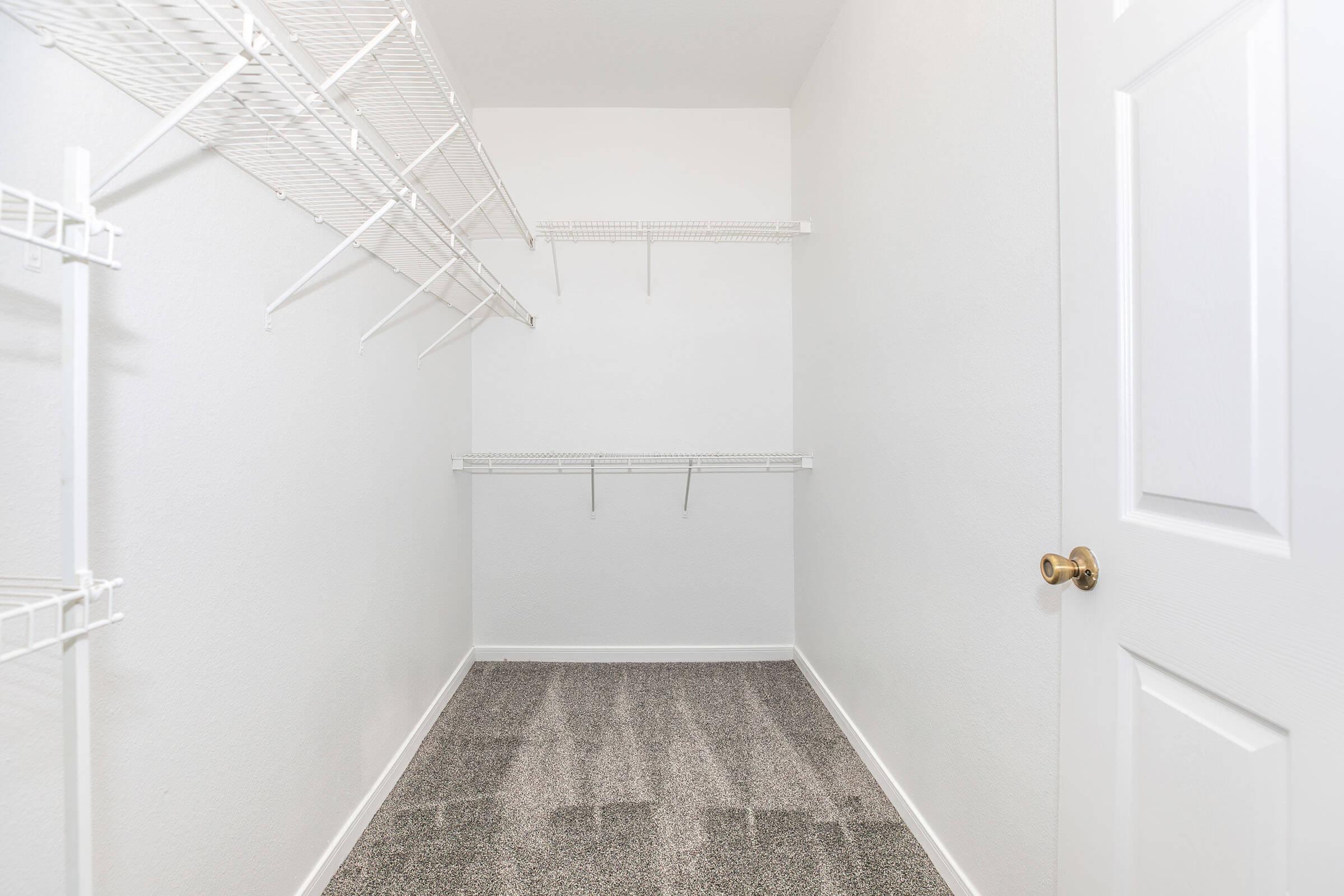 An empty walk-in closet with white walls and a light gray carpet. It features several wire shelves mounted on the walls, providing storage options. The closet has no visible items, creating a clean and spacious appearance, with a closed white door visible on the right side.