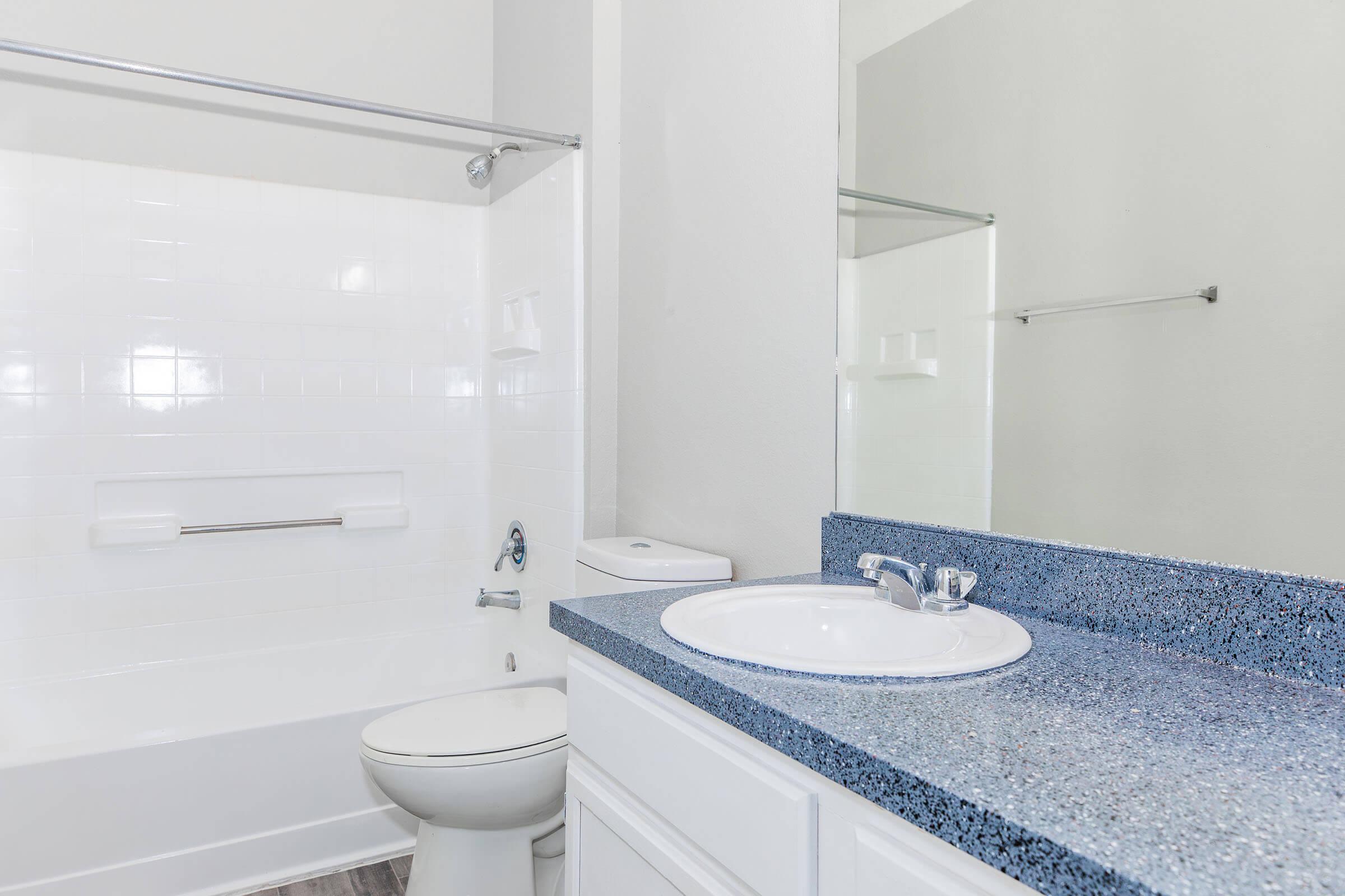 A clean, modern bathroom featuring a white bathtub with a shower, a blue speckled countertop with a sink and faucet, and a white toilet. The walls are painted light gray, and there's a mirror reflecting the space. The overall appearance is bright and airy, with minimal decoration.