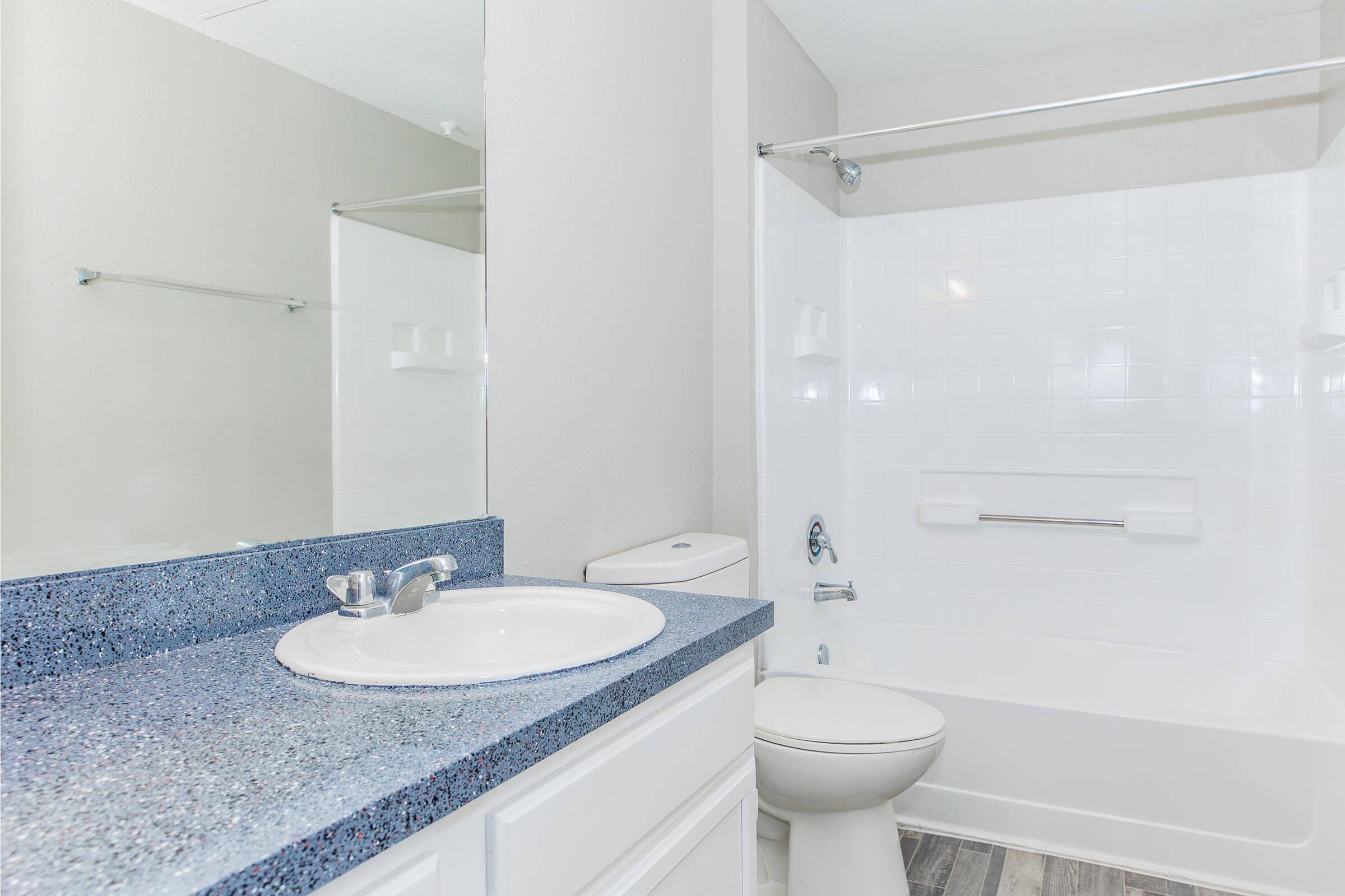 A clean bathroom featuring a white toilet, a bathtub with a shower, a blue speckled countertop with a sink, and a large mirror. The walls are painted in a light color, and there are towel racks and a shower curtain rod visible. The flooring is a light wood-like design.