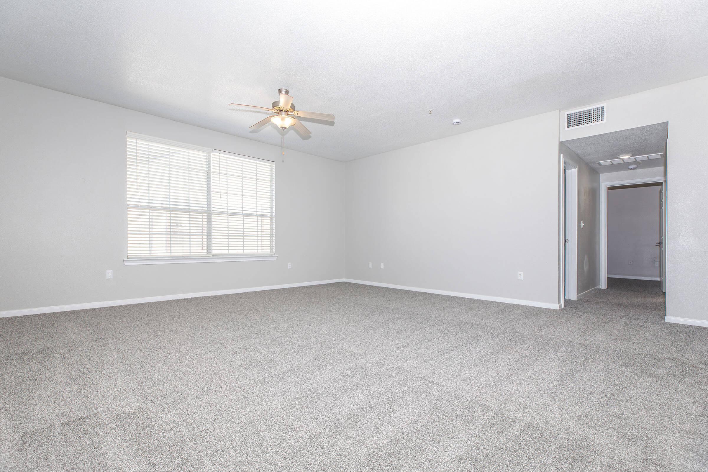A spacious and empty living room featuring light gray carpet, a ceiling fan, and large windows with horizontal blinds. The walls are painted in a neutral tone, creating a bright and airy atmosphere. A doorway leads to another room, adding to the open feel of the space.