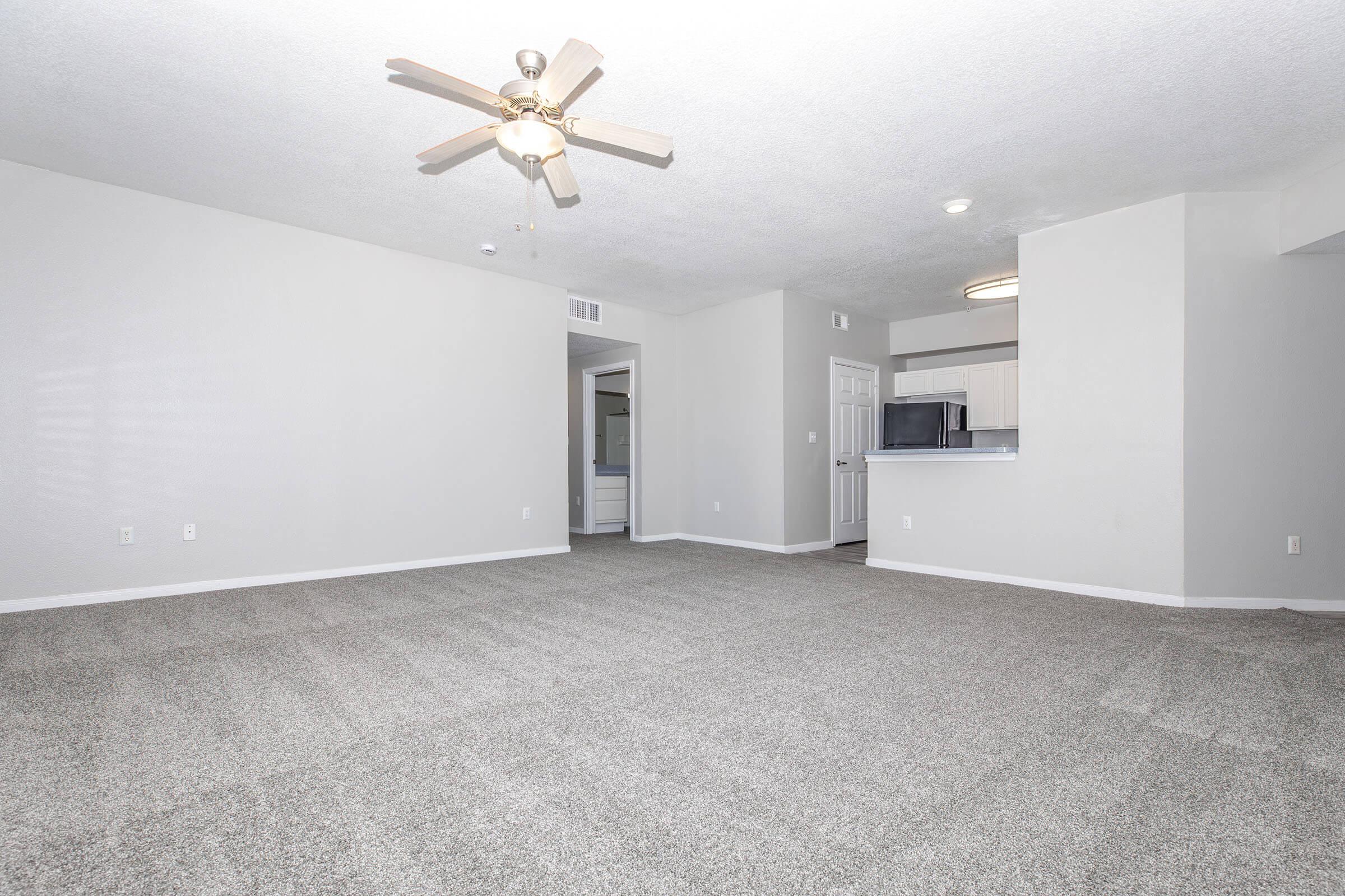 A spacious, well-lit living room featuring light gray walls and plush carpeting. A ceiling fan hangs from the ceiling, and an open doorway leads to another room. In the background, there is a partial view of a kitchen with white cabinets and appliances. The overall atmosphere is modern and inviting.