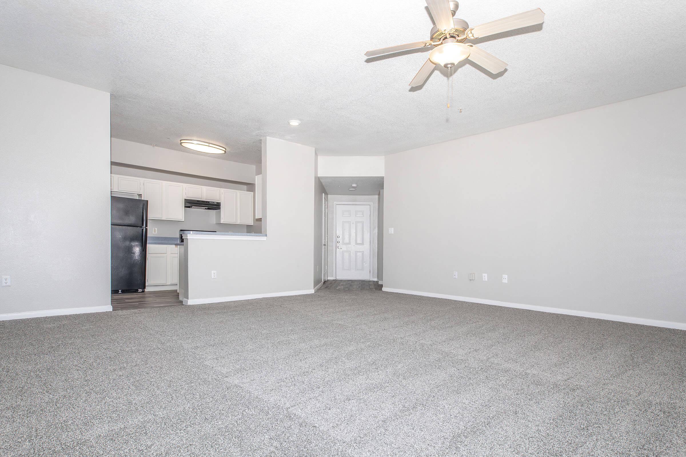 A spacious living area with light gray carpet and a ceiling fan. In the background, there's a kitchen with white cabinetry and black appliances. A doorway leads to another area, and the walls are painted in a neutral color, creating a bright and open atmosphere.