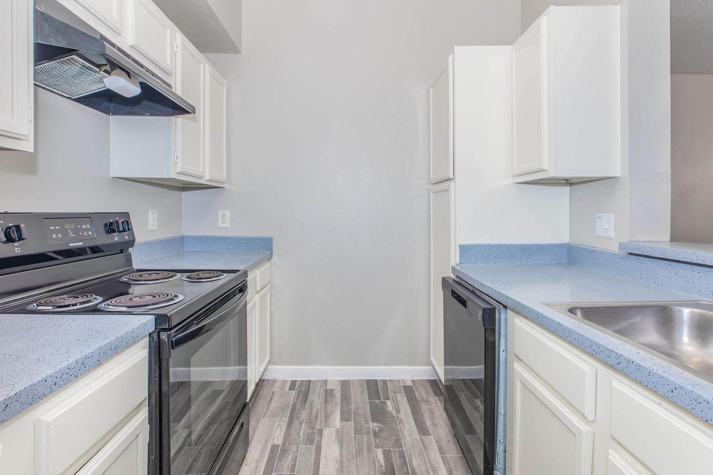A modern kitchen featuring white cabinets, a black stove and oven, and a dual-door refrigerator. The countertops are light blue with a textured finish, and the floor is covered in plank-style tiles. The walls are painted a light gray, creating a contemporary and clean aesthetic.