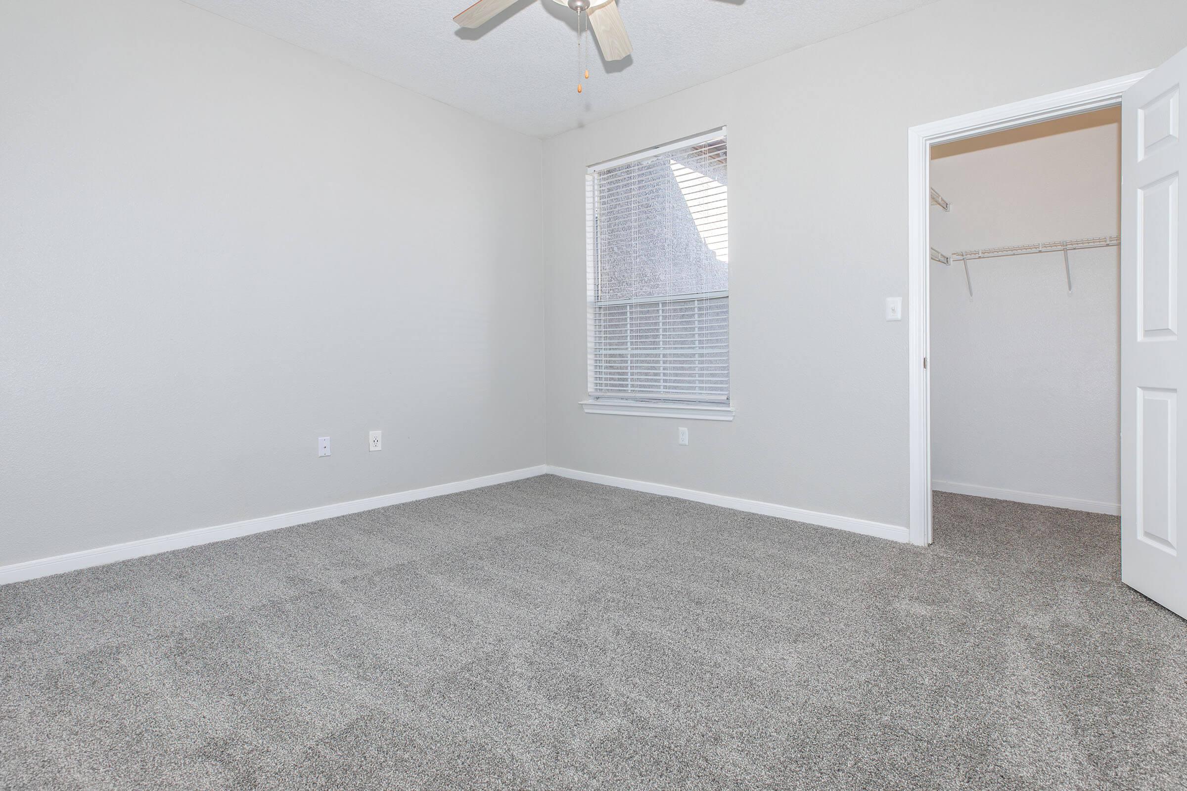 Empty room with light gray walls and new carpet. A ceiling fan is mounted above, and there is a window with blinds letting in natural light. To the right, an open door leads to a closet with a hanging rod and no shelves visible. The overall atmosphere is clean and airy, suitable for various uses.