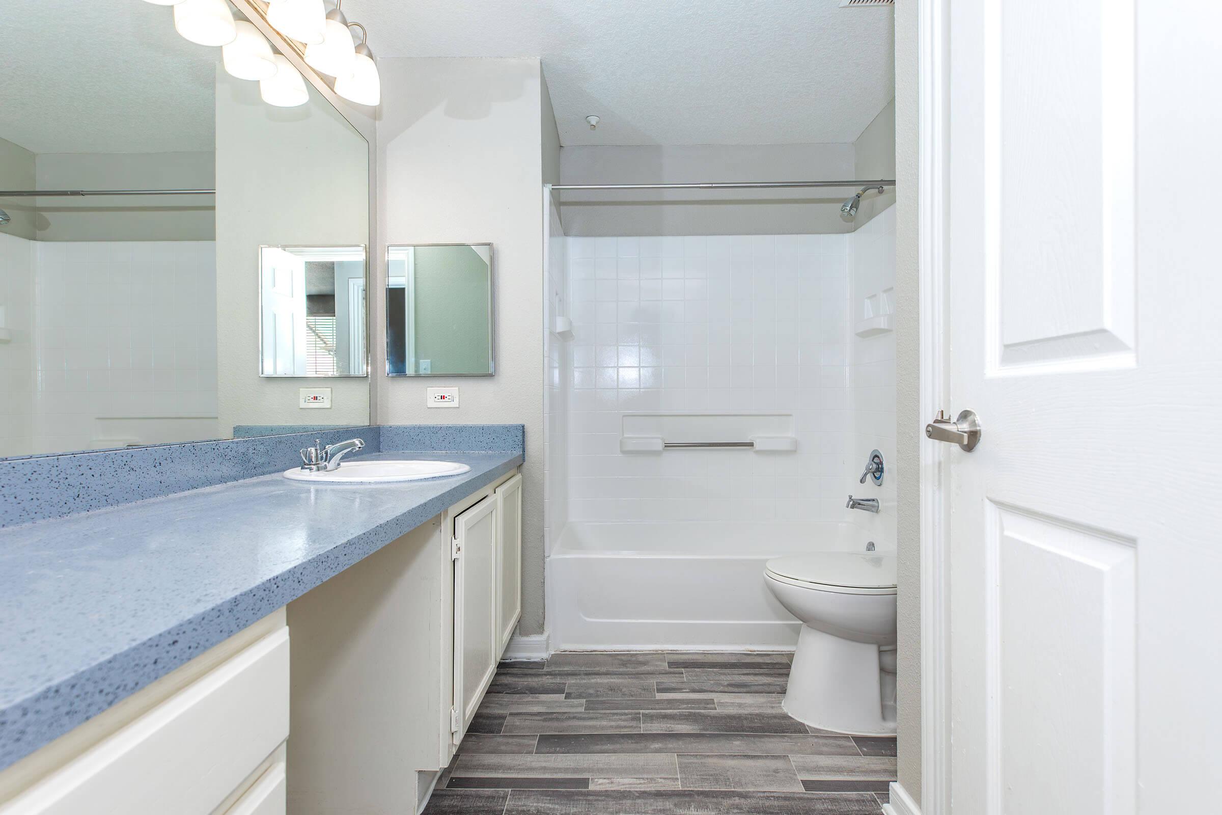 A clean and modern bathroom featuring a light grey countertop with a single sink, a wall-mounted mirror, and a shower-tub combo. The space has dark vinyl flooring and a white door leading to the hallway. Bright lighting fixtures are installed above the mirrors.