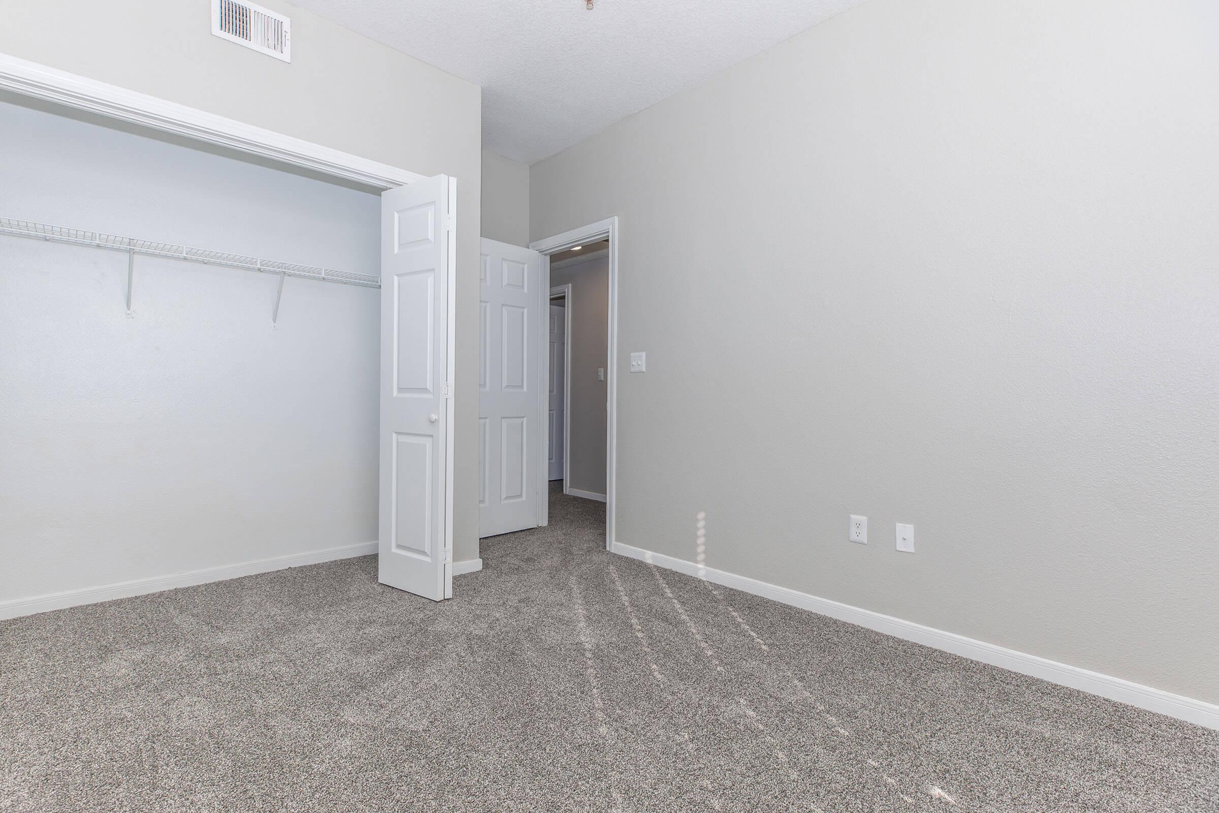 Empty room with light gray walls and freshly carpeted floor. Two open doors, one leading to a closet with a shelf, and the other to a hallway. Sunlight streaming in through the doorway, creating a bright and airy feel. No furnishings present, highlighting the spaciousness of the area.