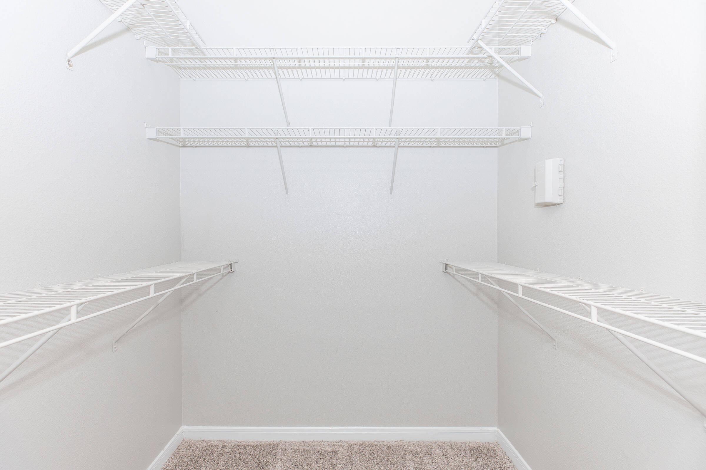 Empty closet with white wire shelves along the walls, featuring a light gray wall and a carpeted floor. The space appears clean and minimalist, with no items visible on the shelves.