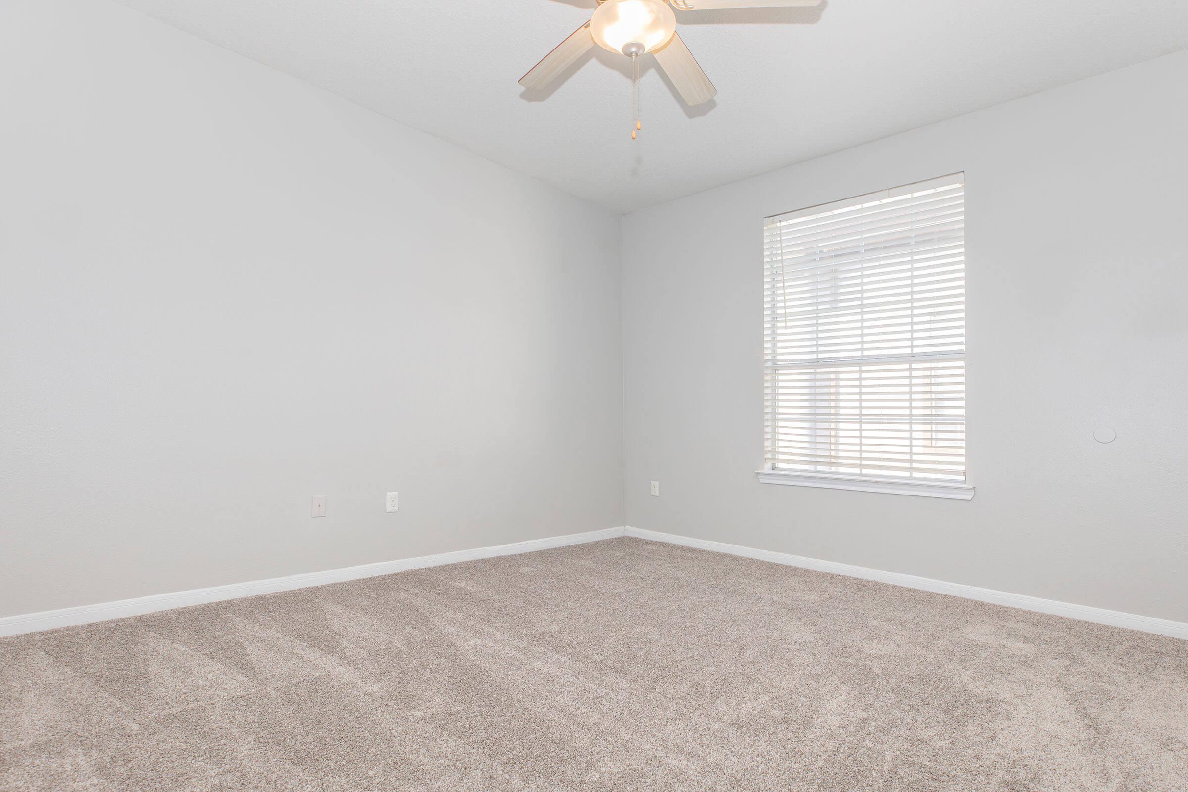 Empty room with light gray walls and a light colored carpet. A ceiling fan hangs from the ceiling, and natural light streams in through a window covered with white blinds. The room has no furniture and appears spacious and bright.