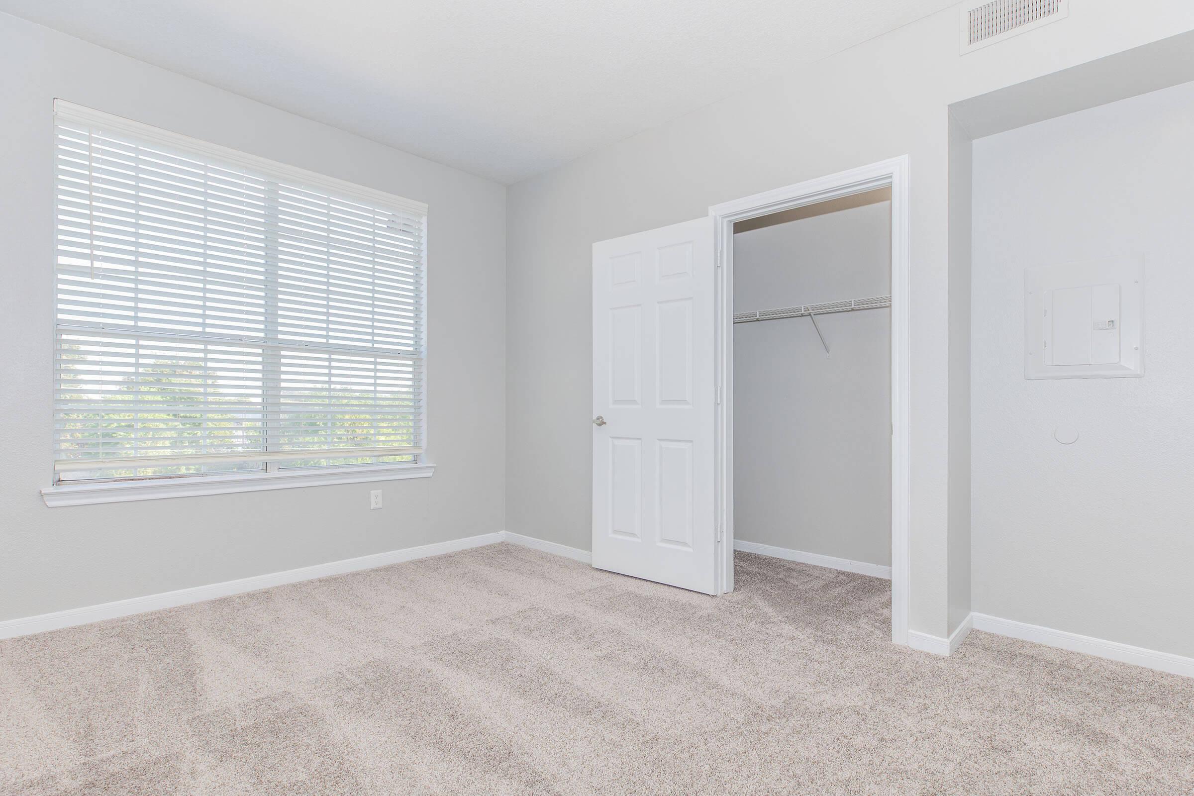 A spacious, empty bedroom featuring light gray walls and plush beige carpet. The room has a large window with blinds allowing natural light in, and a closet with a sliding white door. There is a white electrical panel on the wall, and the overall atmosphere is bright and inviting.