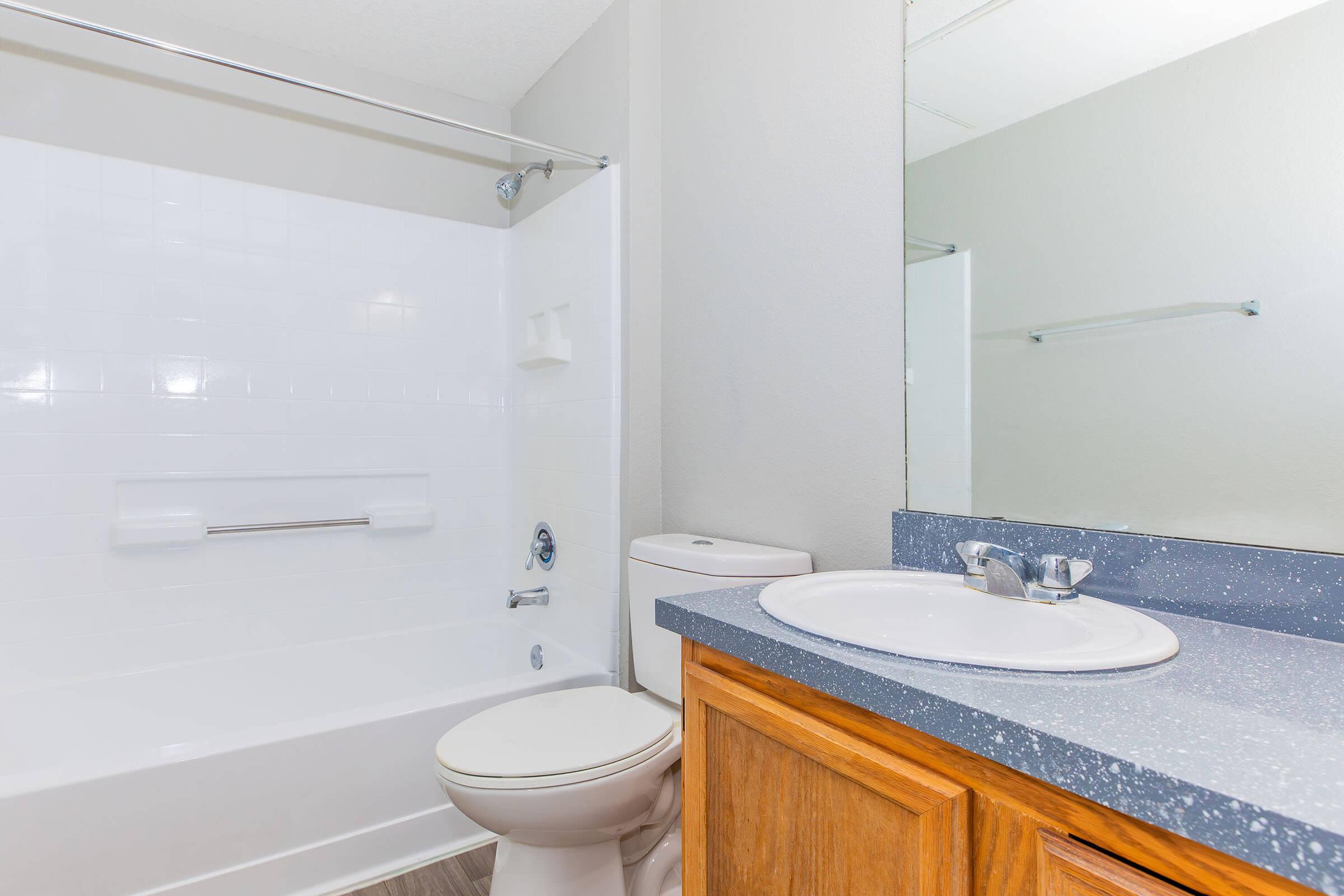 A clean and modern bathroom featuring a white bathtub with a shower, a toilet, a sink with a faucet, and wooden cabinetry. The walls are painted light grey, and there is a large mirror above the sink. Bright, neutral colors create a spacious and inviting atmosphere.