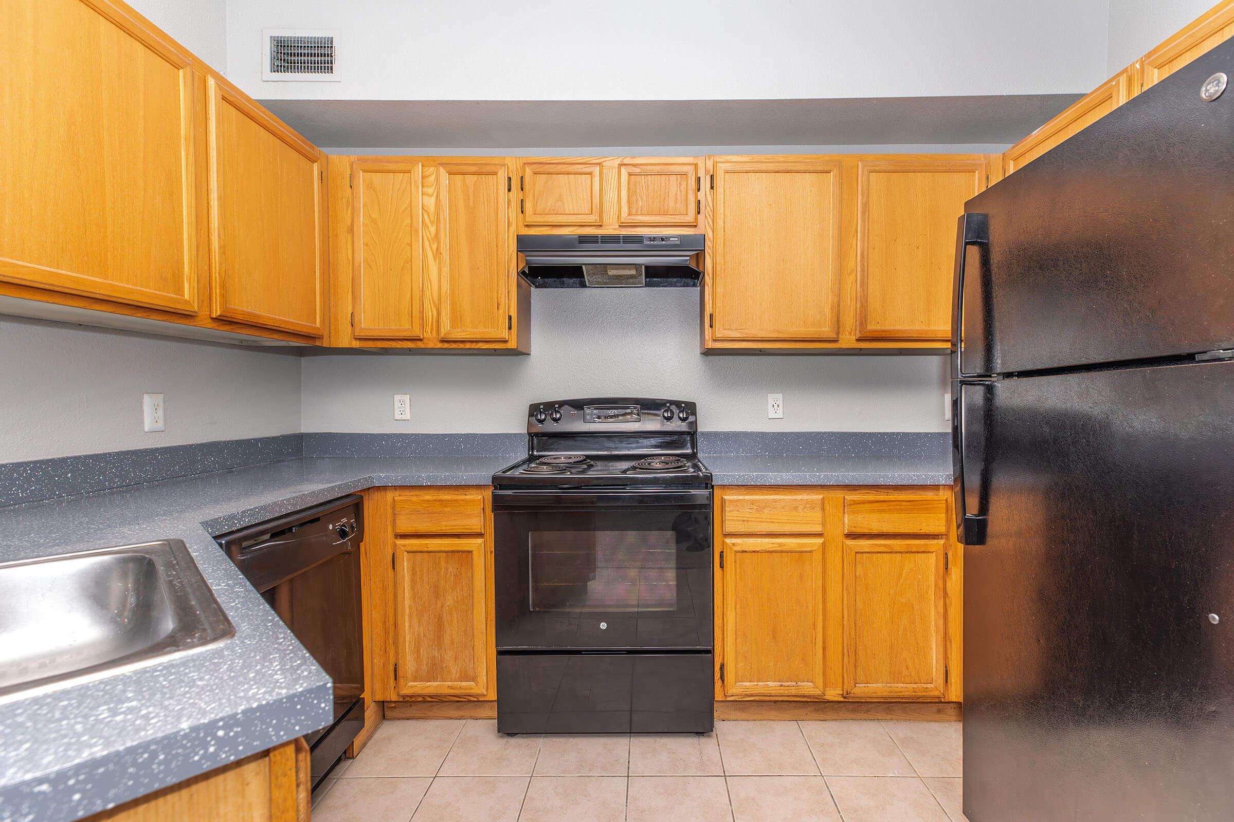 A kitchen featuring wooden cabinets, a stainless steel sink, a black refrigerator, an oven with a range, and a vent hood. The countertops are dark, and the tile flooring is light-colored. Overall, the space is functional and designed for cooking and food preparation.