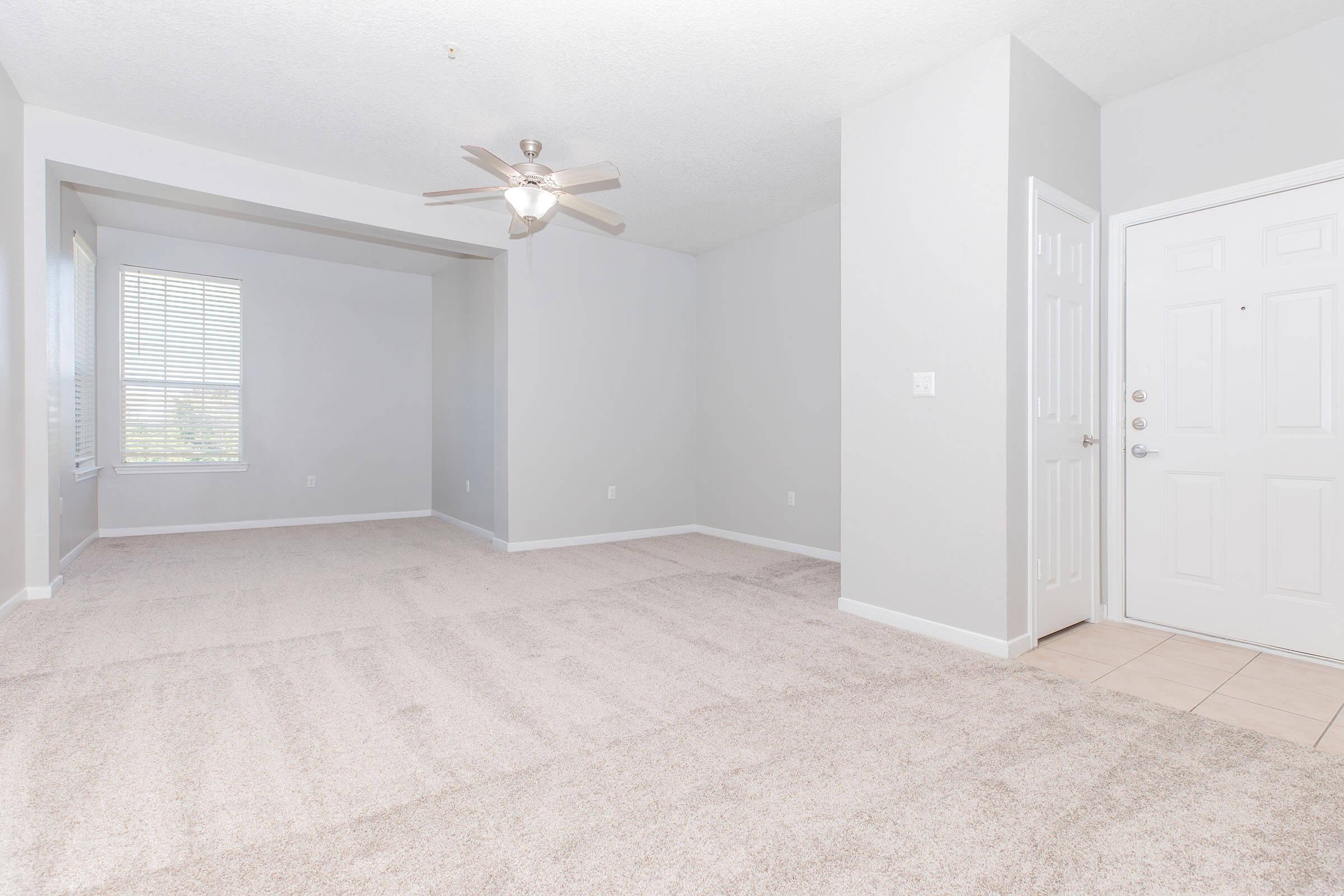 Spacious, empty living room with light gray walls and plush beige carpet. Features a ceiling fan, a large window allowing natural light, and a doorway leading to an entrance area. The space is clean and ready for furnishings, offering a bright and inviting atmosphere.