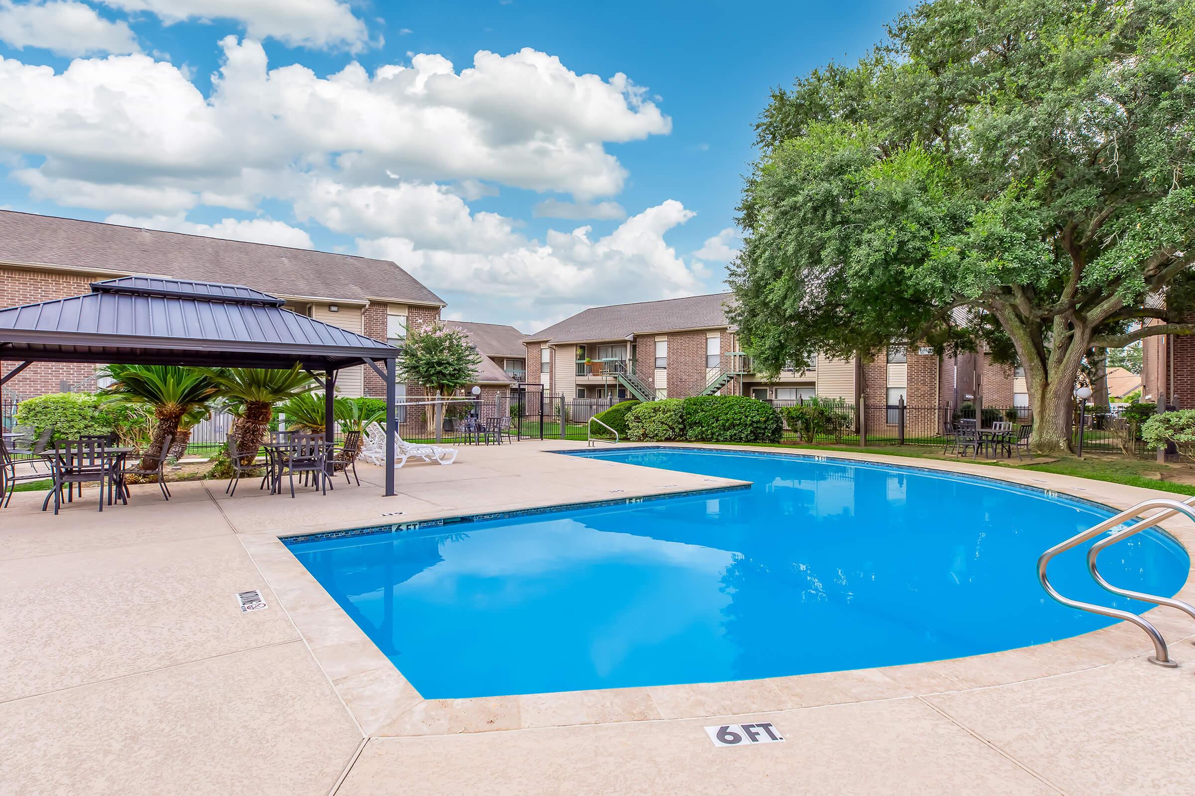 A bright and inviting swimming pool surrounded by lush greenery and palm trees. A shaded gazebo and outdoor seating area can be seen nearby, with residential buildings in the background under a partly cloudy sky. The area invites relaxation and leisure.