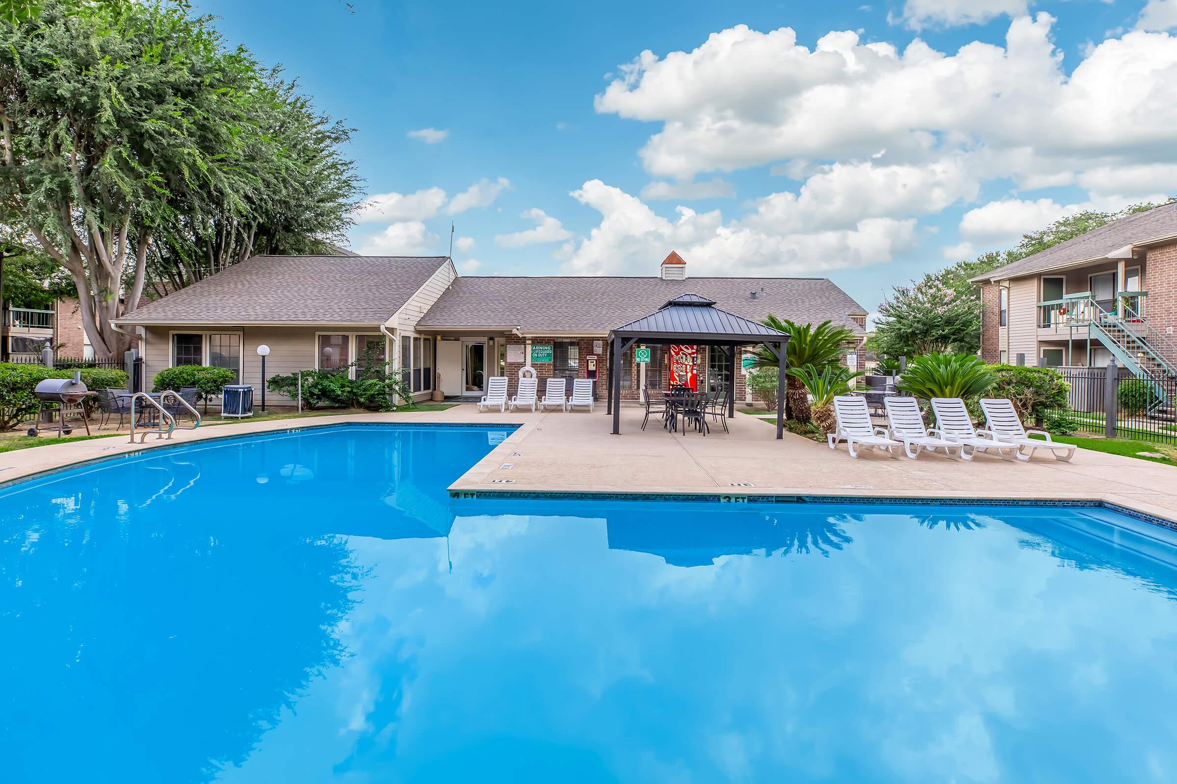 A clear blue swimming pool surrounded by lounge chairs and a gazebo. In the background, there are buildings with balconies and lush greenery. The sky is bright with fluffy white clouds, creating a welcoming atmosphere for relaxation and leisure.